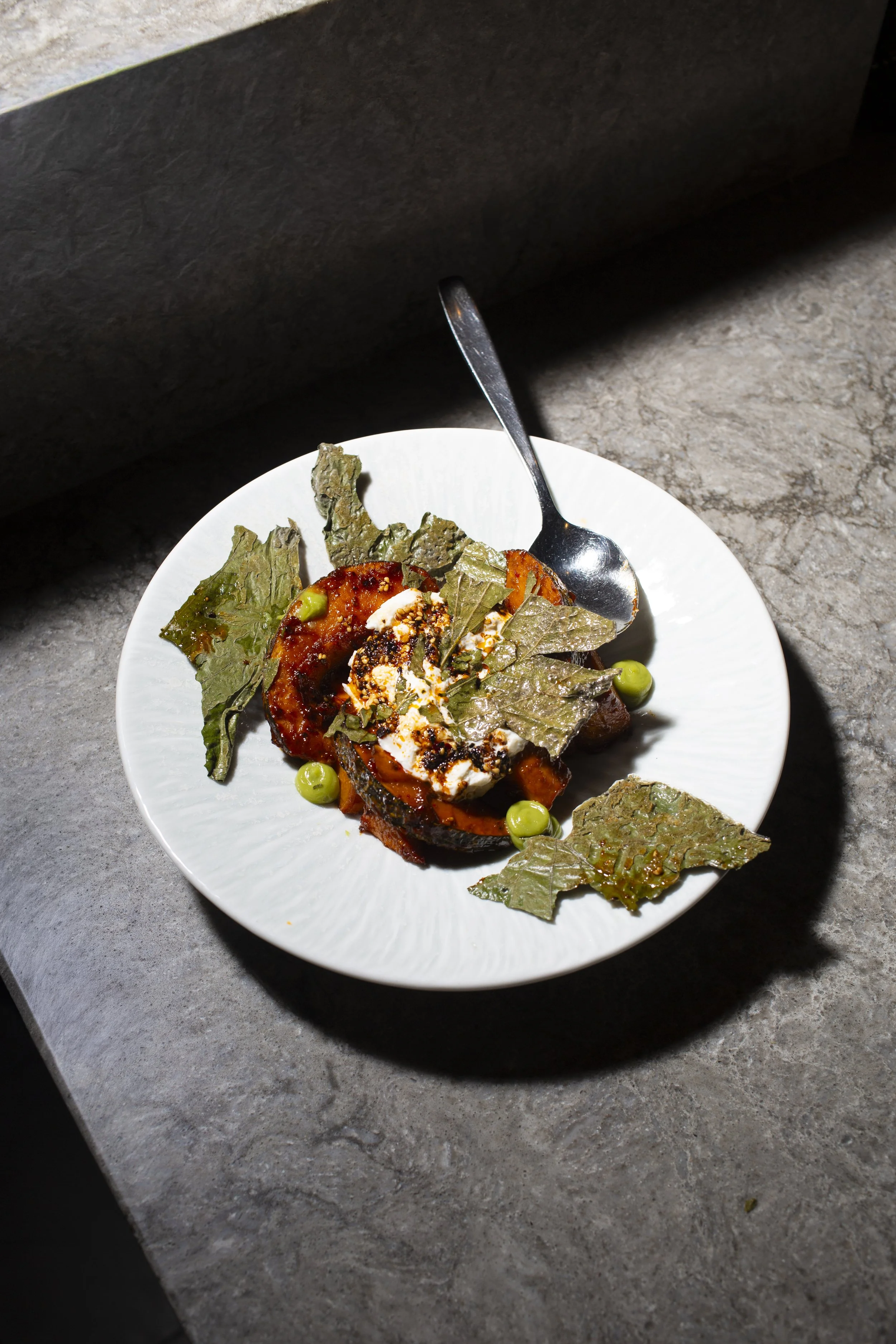 A white plate with grilled vegetables, green peas, and fresh green leaves on a gray stone countertop.