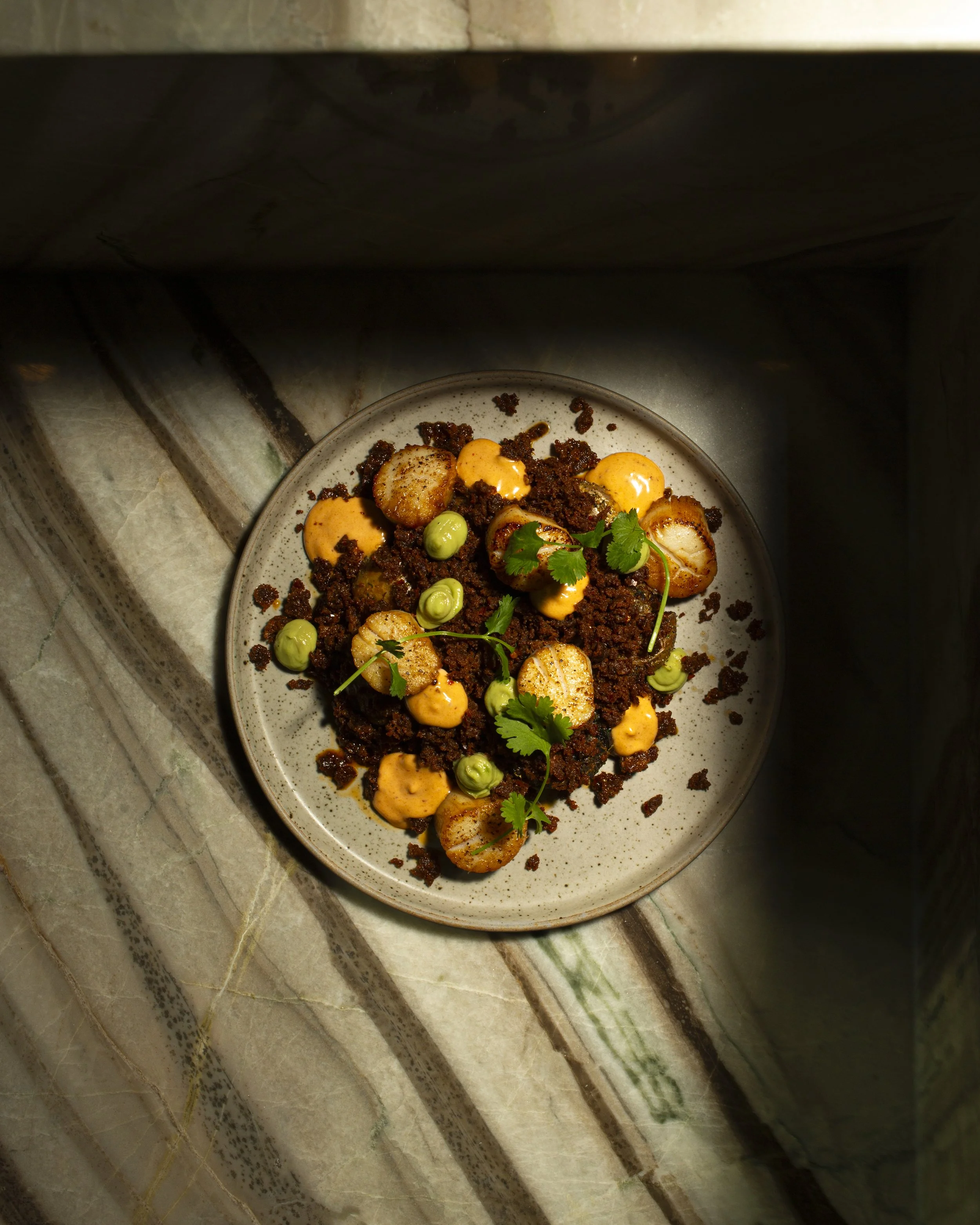 A plate of food with scallops, ground meat, green peas, and cilantro garnished on a beige speckled plate. The plate is on a stone surface.