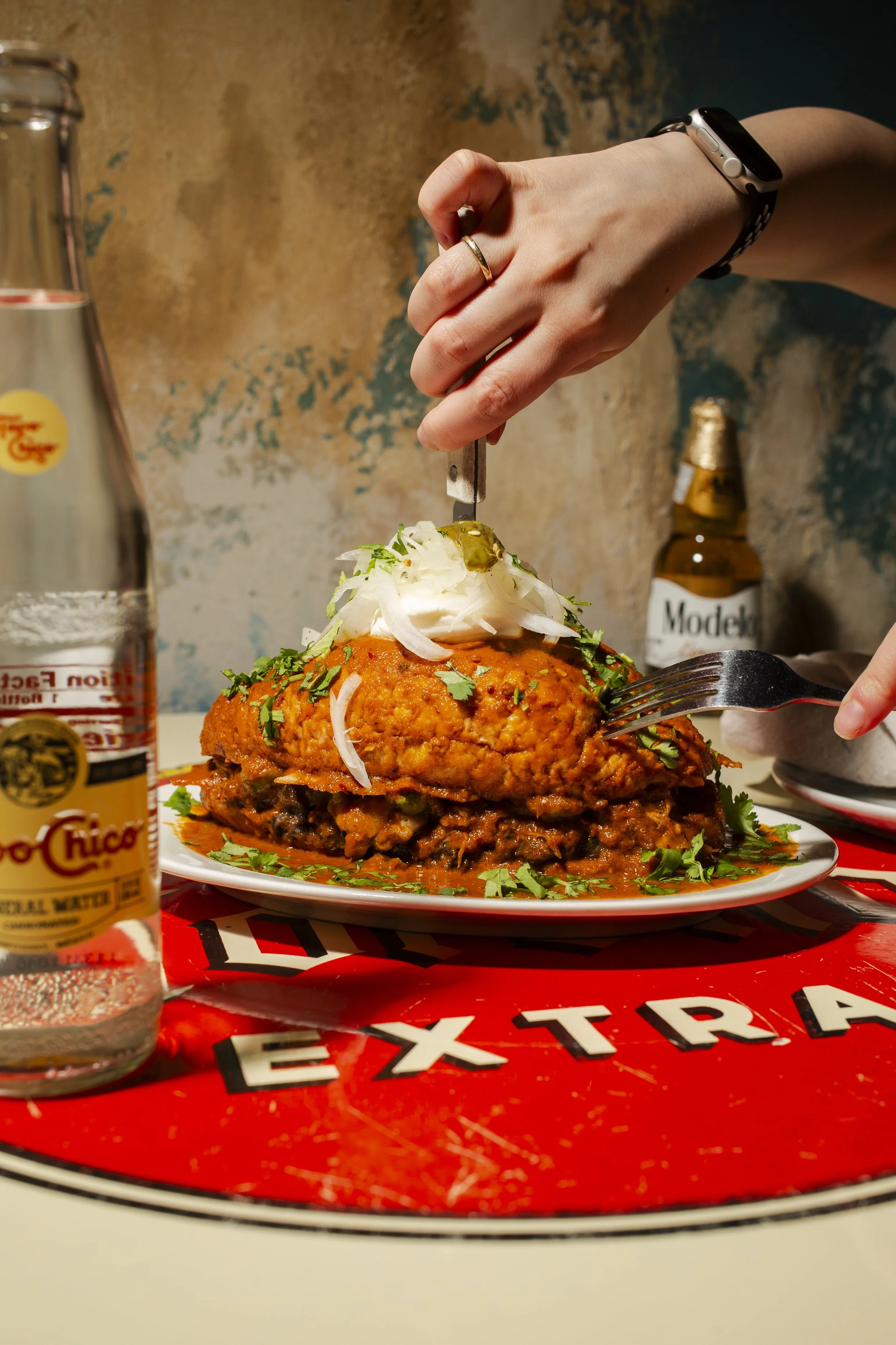 A plate with a large serving of Mexican food, topped with sour cream, onions, cilantro, and jalapeños, with a fork and knife, on a table with a red placemat that says 'EXTRA'. A glass bottle of Mexican soda, a small beer bottle, and a bowl are on the