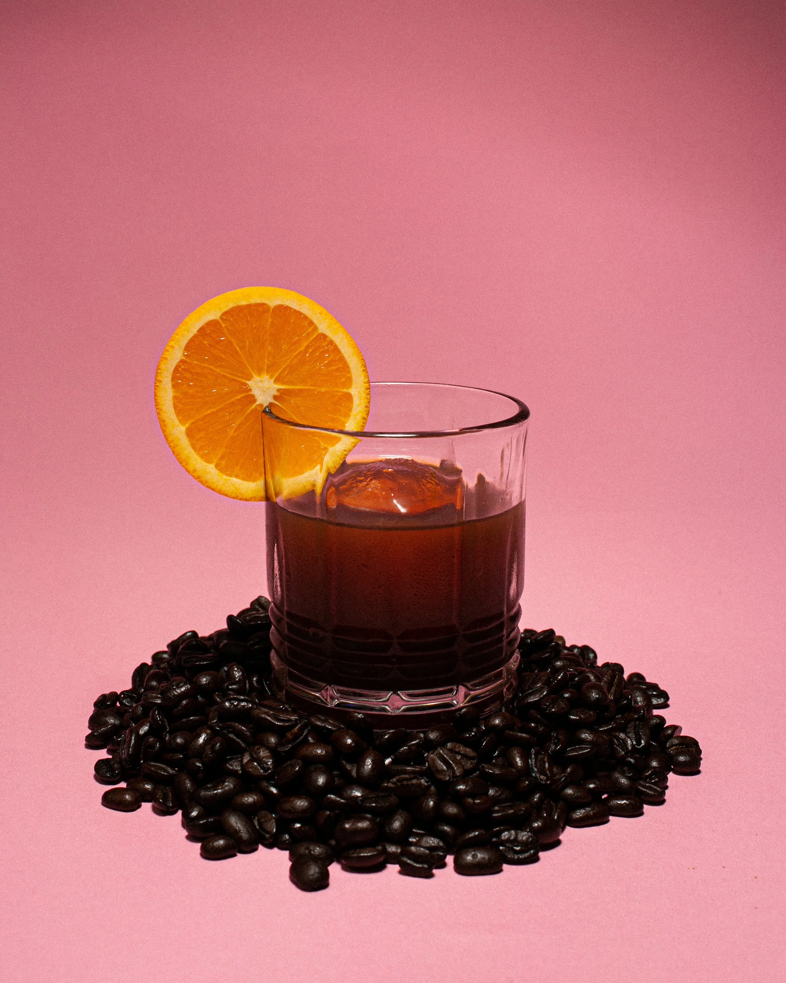 A glass of dark coffee with ice, garnished with a slice of orange on the rim, sitting on a pile of coffee beans against a pink background.