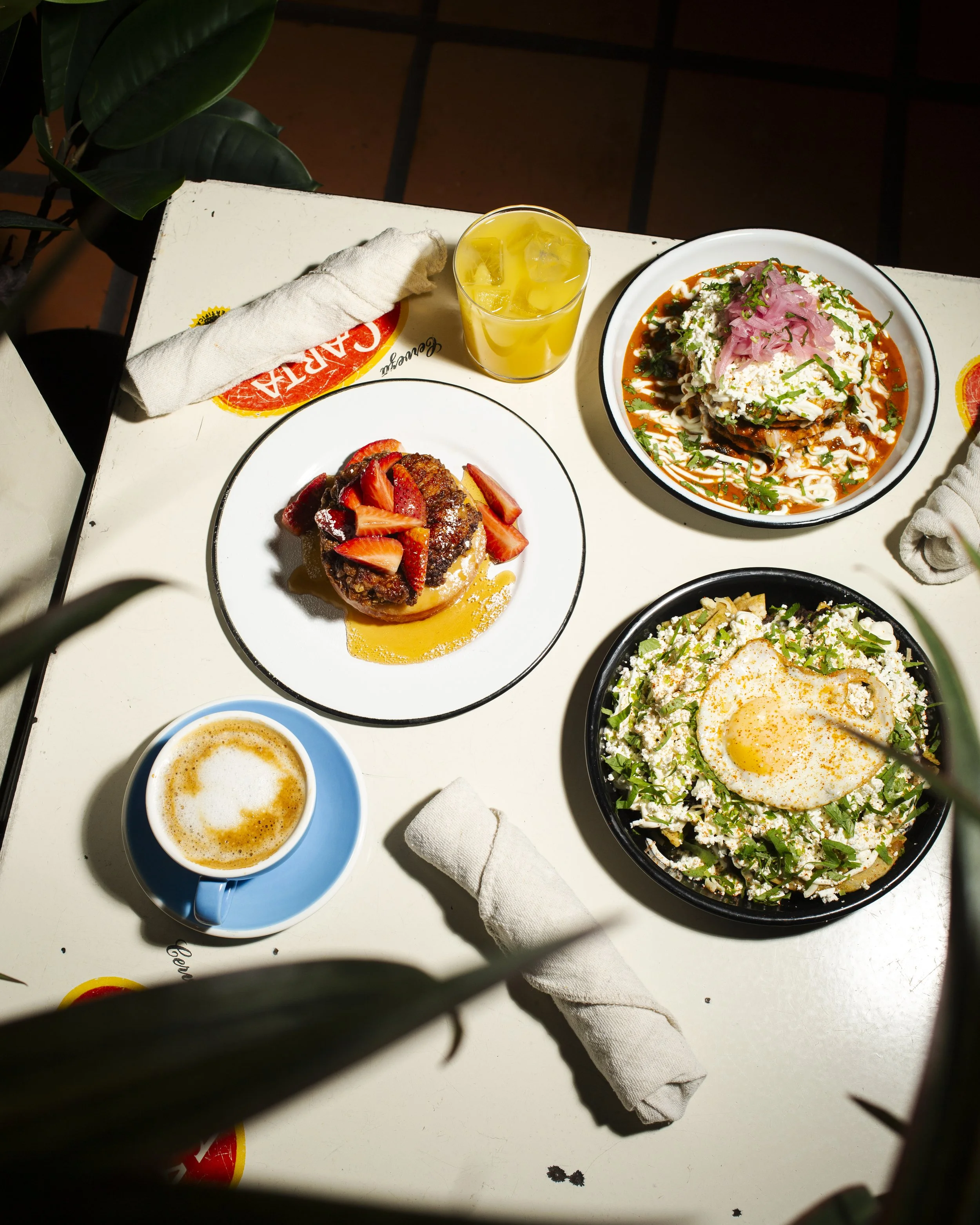 Top-down view of a table with various dishes including a cup of coffee, a glass of iced yellow beverage, a plate of a dessert with strawberries, a bowl of salad with an egg on top, and a plate of Mexican food with toppings.