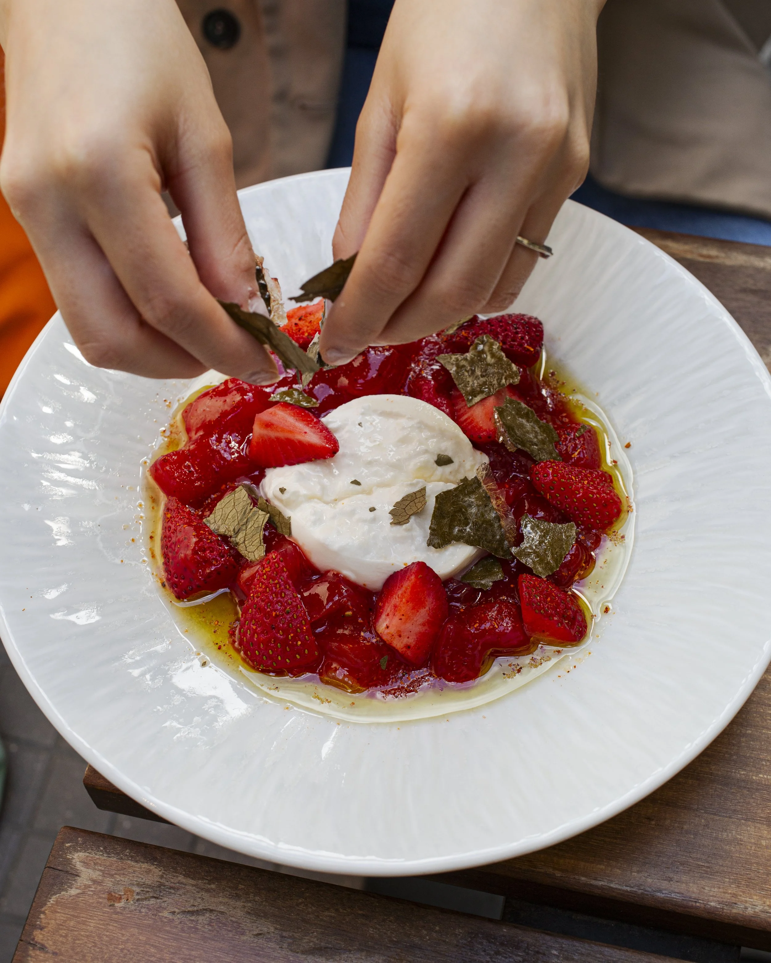 A person garnishing a strawberry dessert with basil leaves in a white bowl.