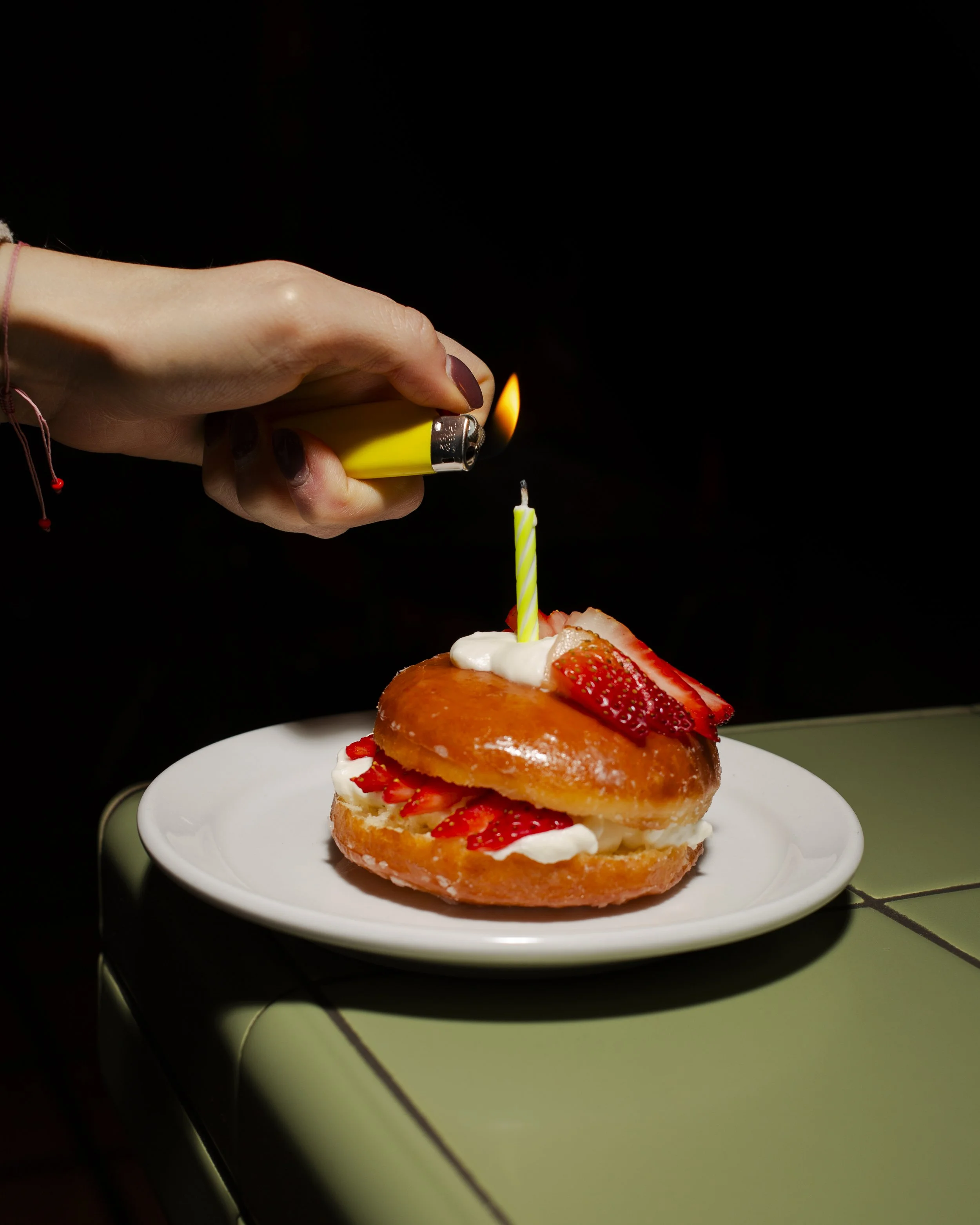 A hand holding a yellow lighter is lighting a birthday candle on a strawberry-filled pastry. The pastry has strawberries, whipped cream, and a slice of strawberry on top, placed on a white plate on top of a green tiled surface.