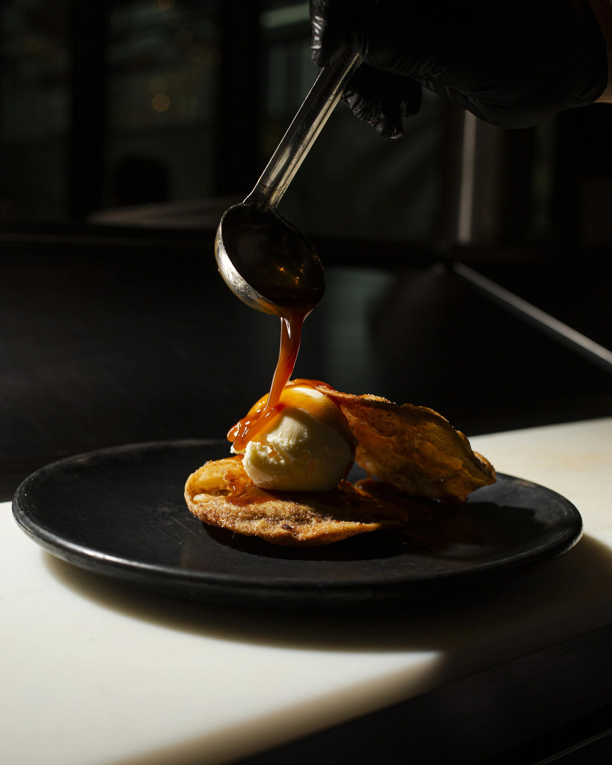 Scoop of vanilla ice cream on a crispy waffle with caramel sauce being poured over it on a black plate.