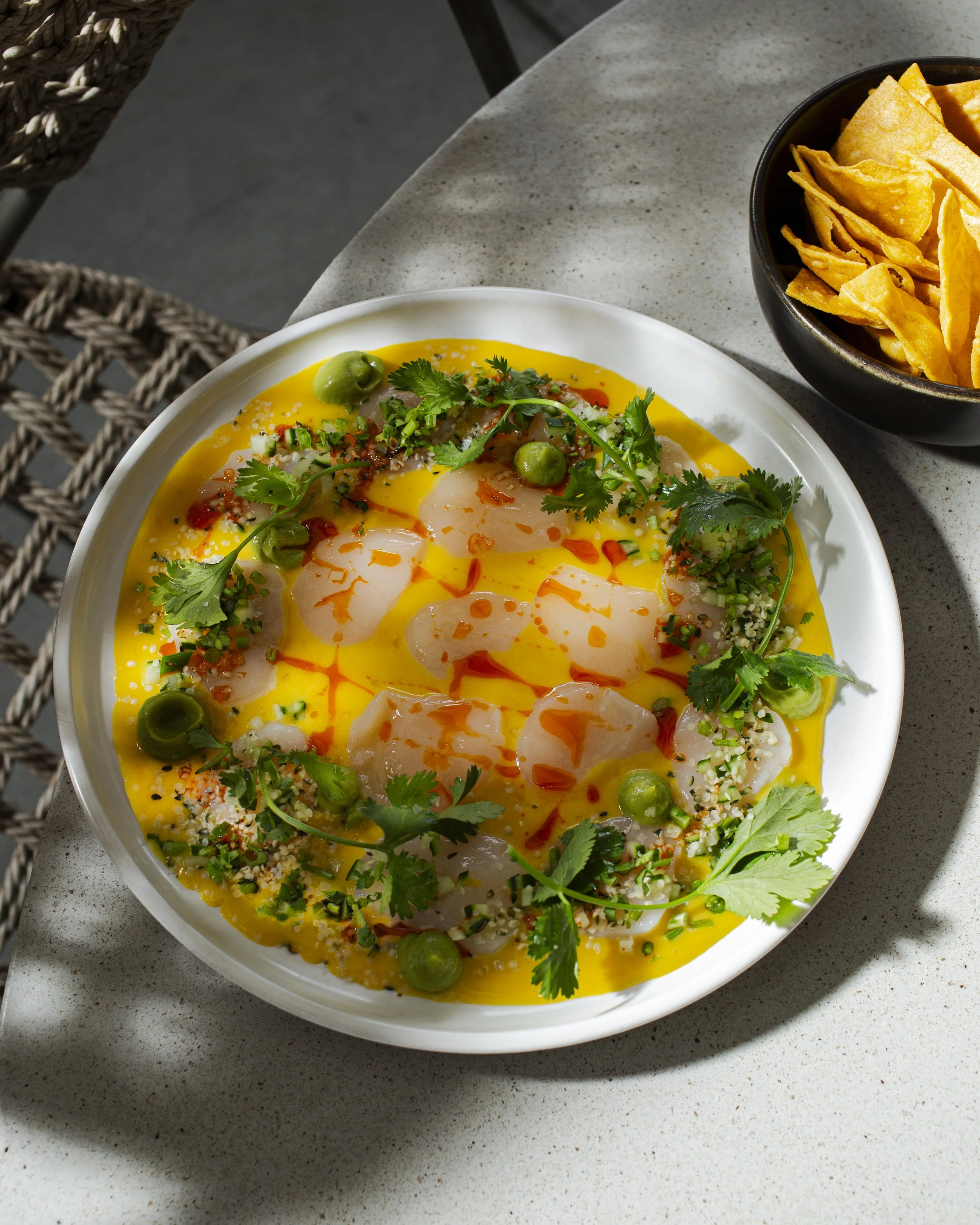 A dish of yellow sauce with raw fish slices, green peas, cilantro, and red chili oil, garnished with herbs and seeds, served with a side of tortilla chips.