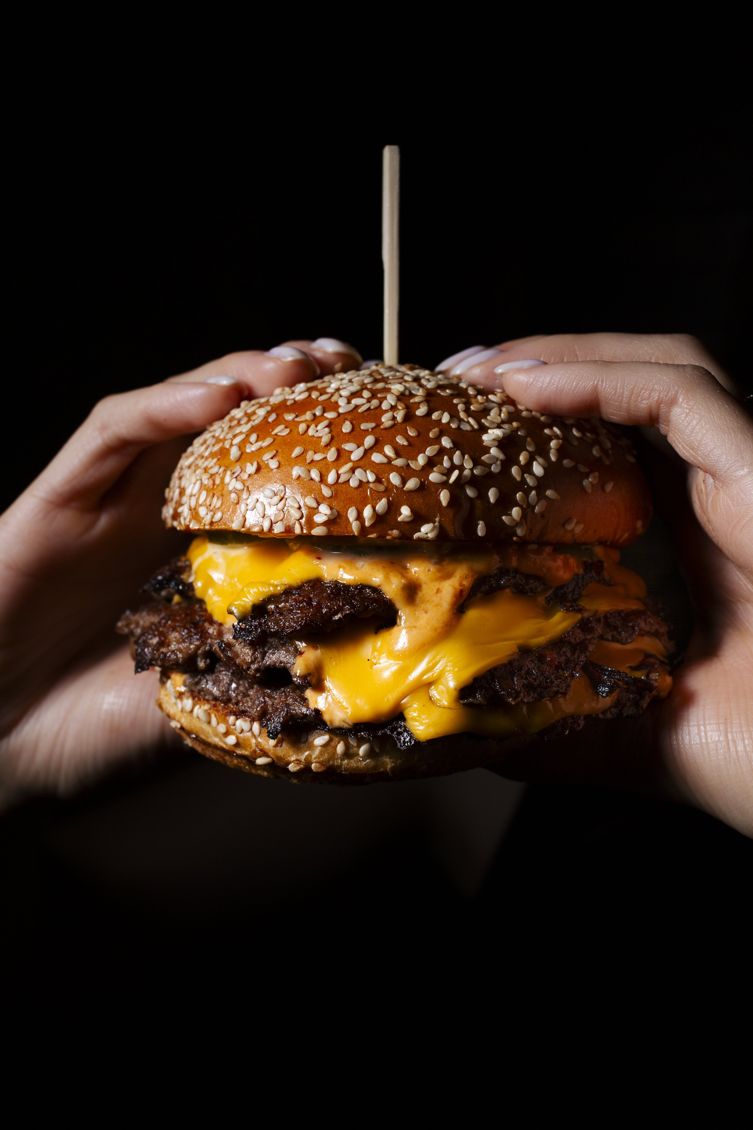 Close-up of a person holding a cheeseburger with hands on a black background. The burger has a sesame seed bun, melted cheddar cheese, beef patties, and a wooden skewer on top.