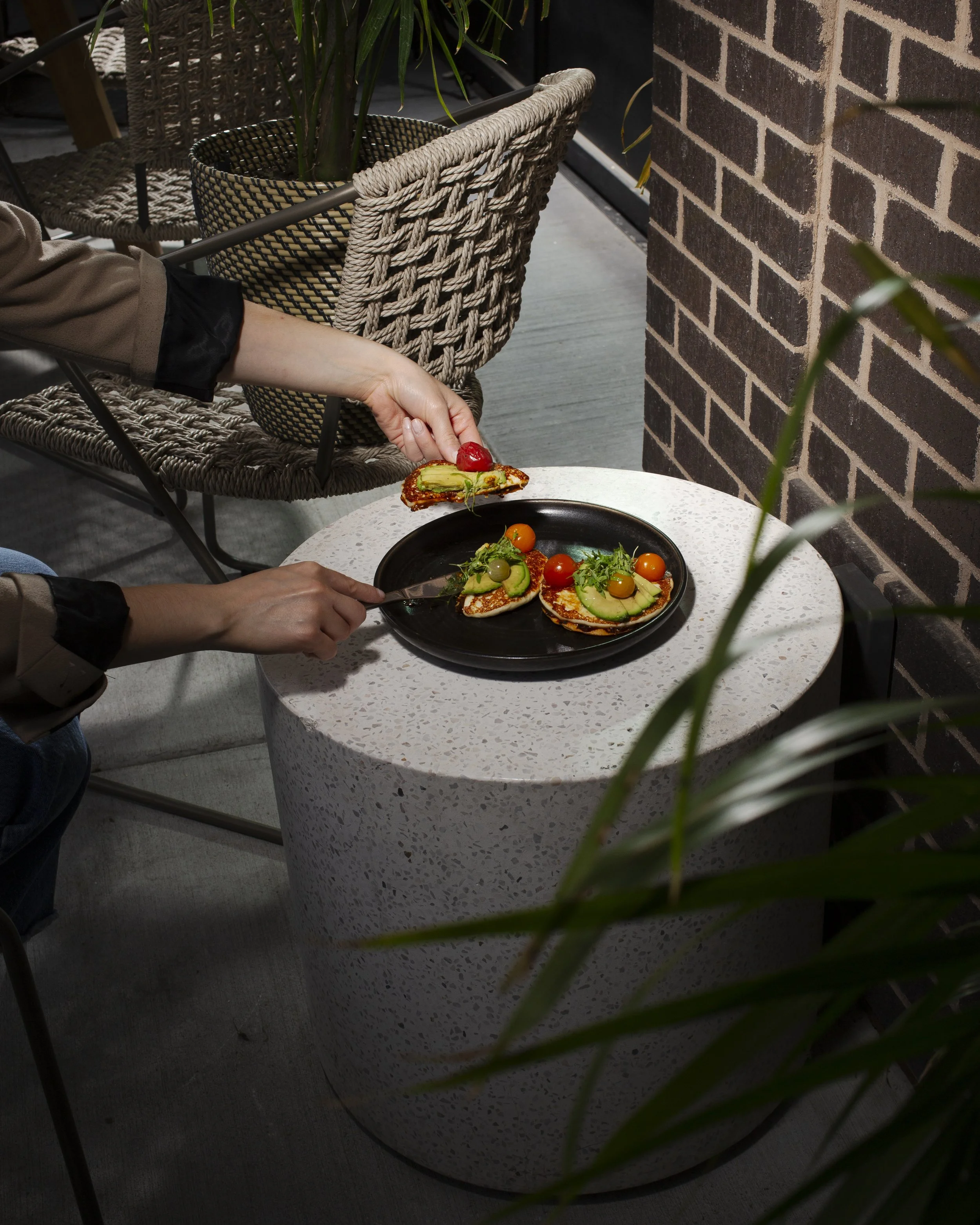 Person serving colorful vegetable dishes, including cherry tomatoes and avocado slices, onto a black plate on a white speckled table in a cozy indoor setting with brick wall and potted plants.