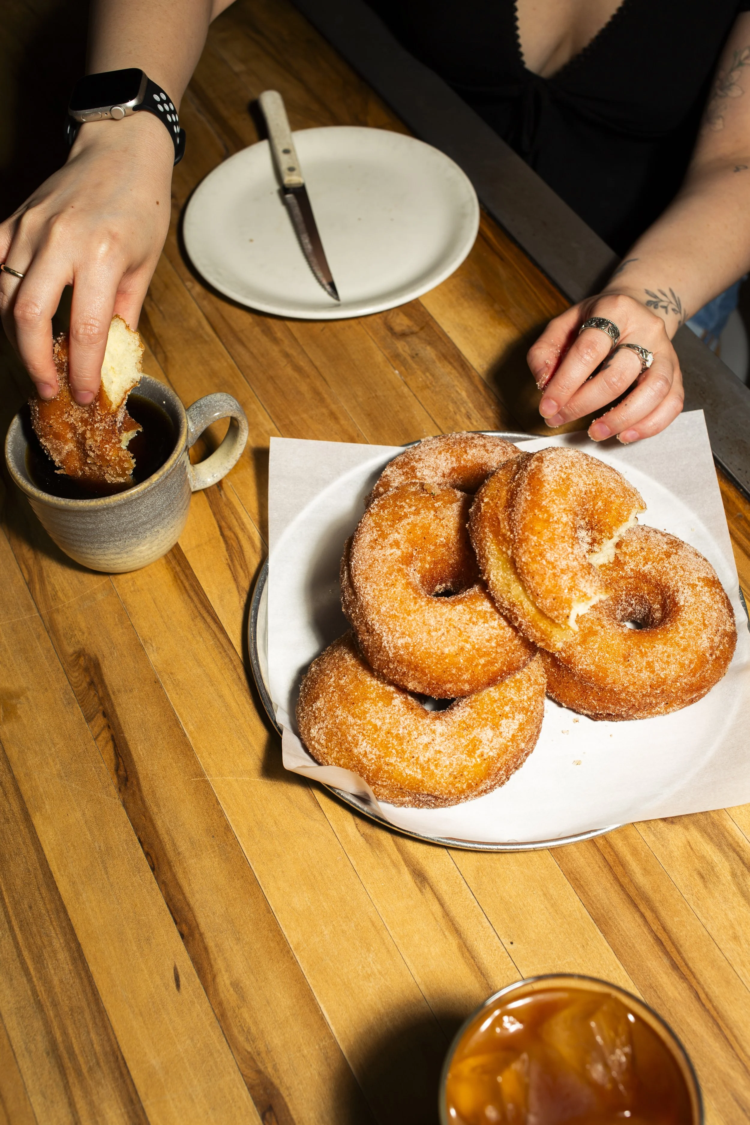 Four sugar-coated donuts on a white plate on a wooden table, with a person dipping a donut into a mug of coffee or hot chocolate, wearing rings and a smartwatch.