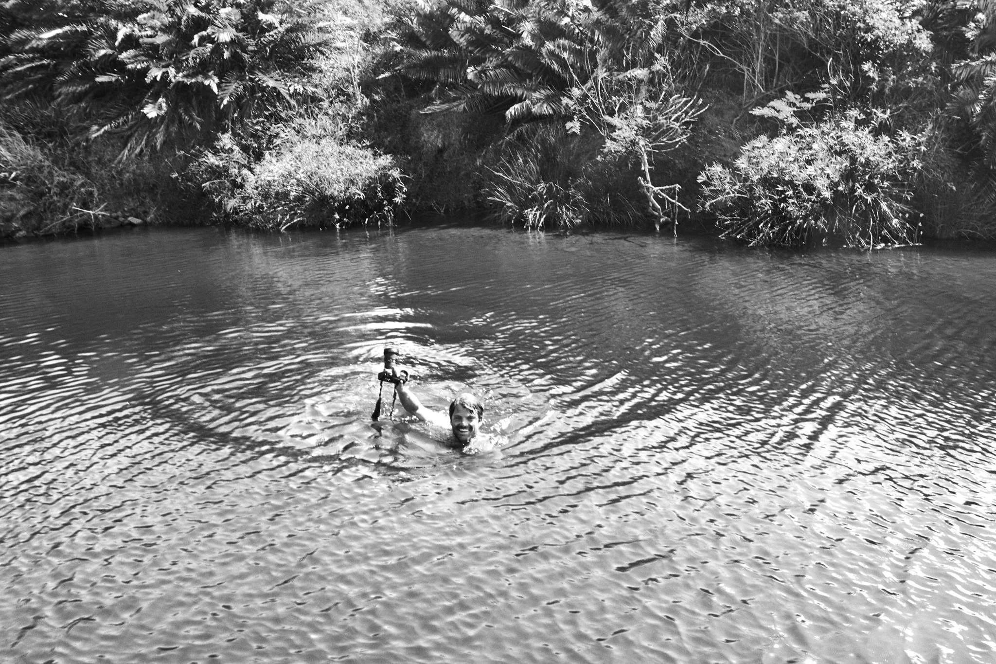 A man swimming in a river, holding a camera in his right hand, surrounded by lush vegetation.