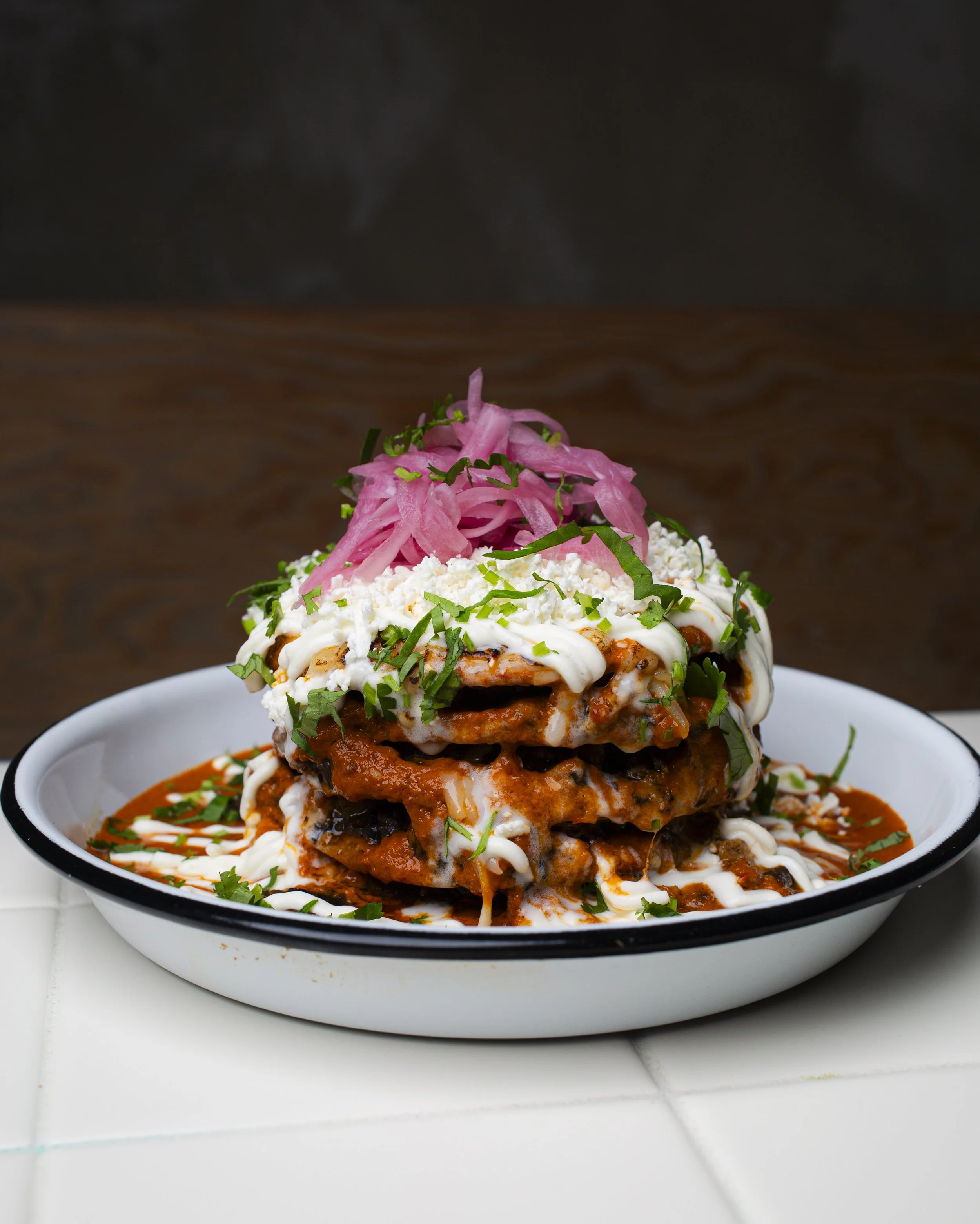 A plate of layered Mexican enchiladas topped with cheese, sour cream, chopped cilantro, and pickled red onions, with sauce drizzled around the edges.