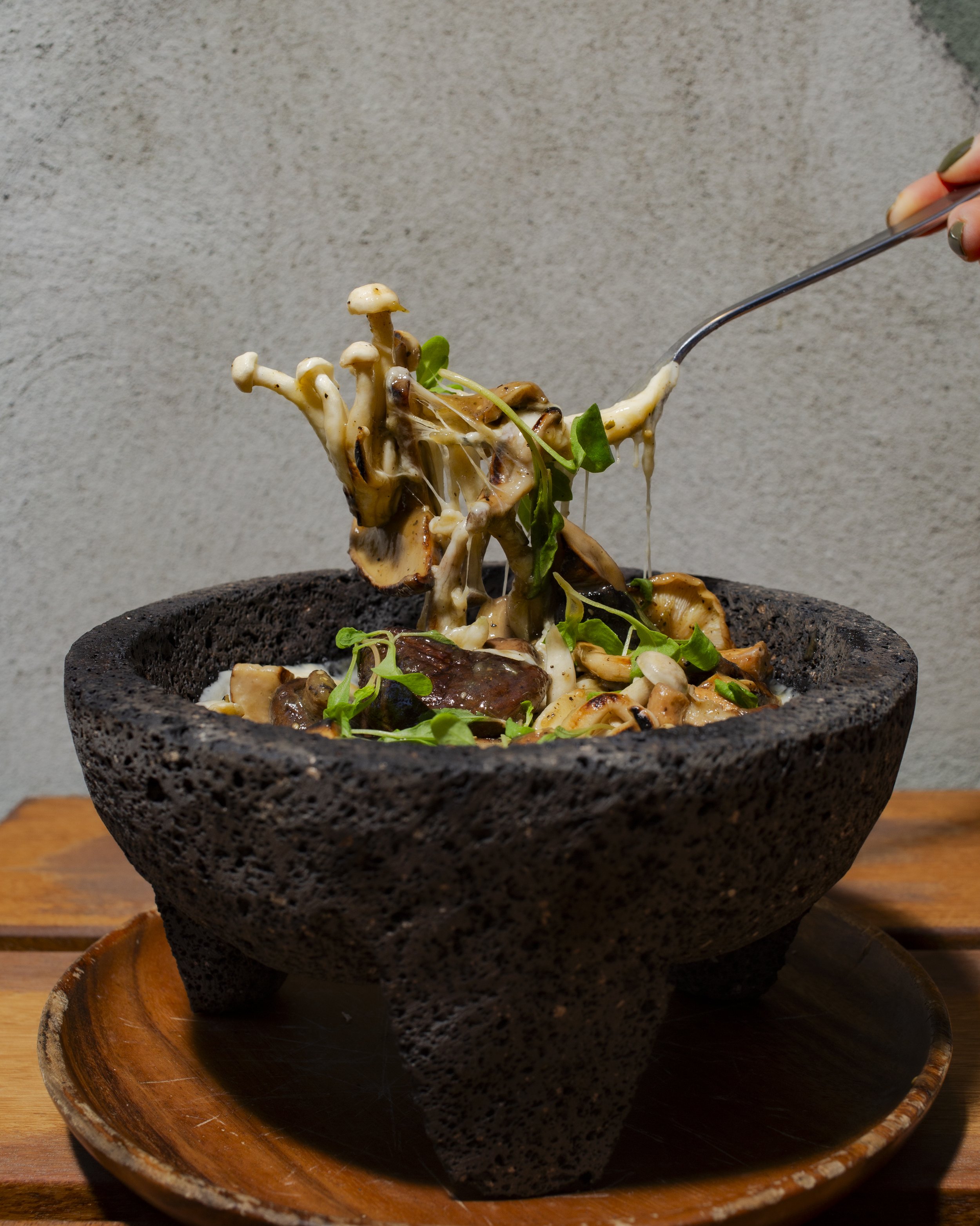 A person lifting a spoonful of mushroom and greens salad from a stone bowl on a wooden tray.