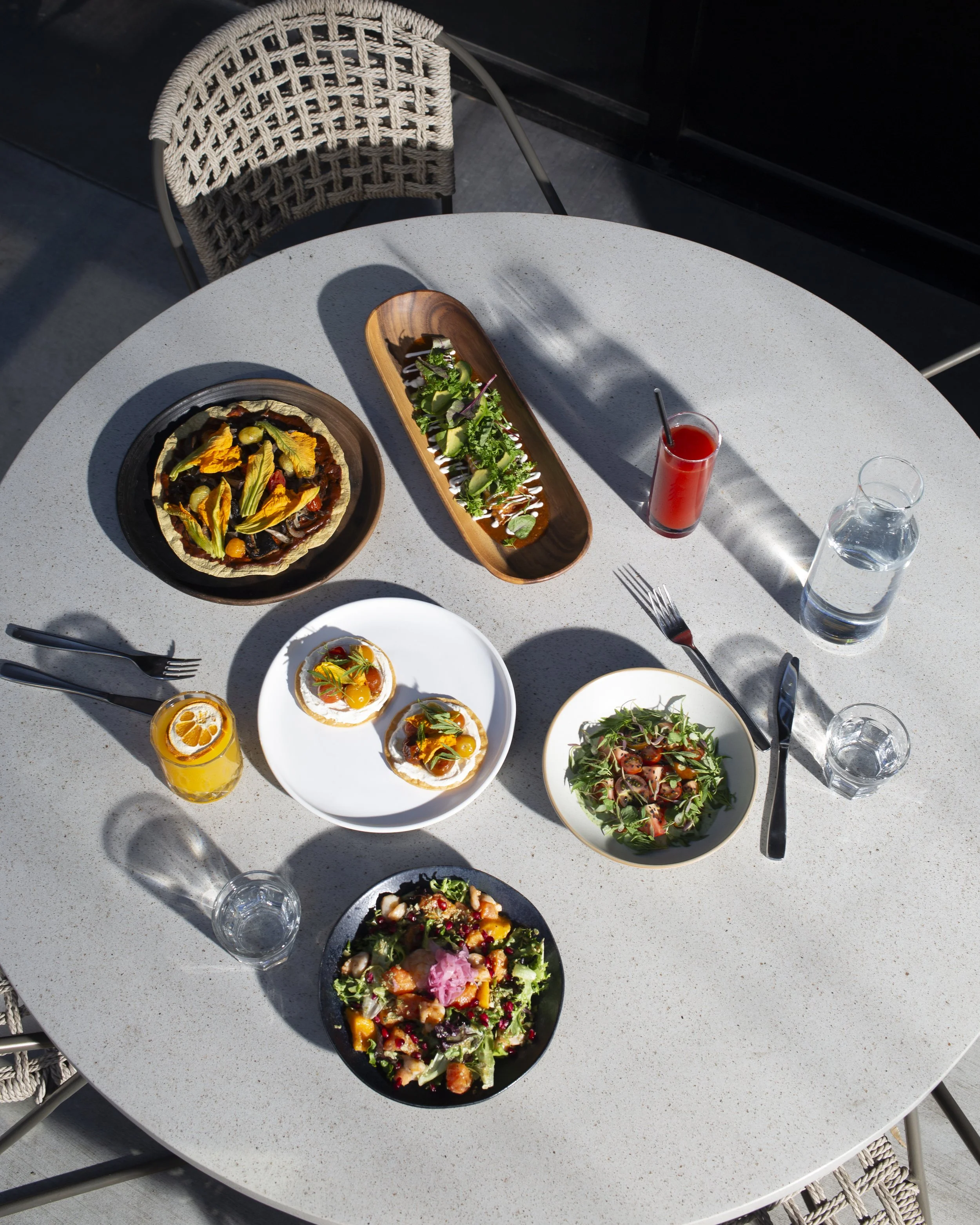 Overhead view of a round table with various dishes including salads, a sourdough pizza, and drinks in glasses, with two empty water glasses and cutlery.