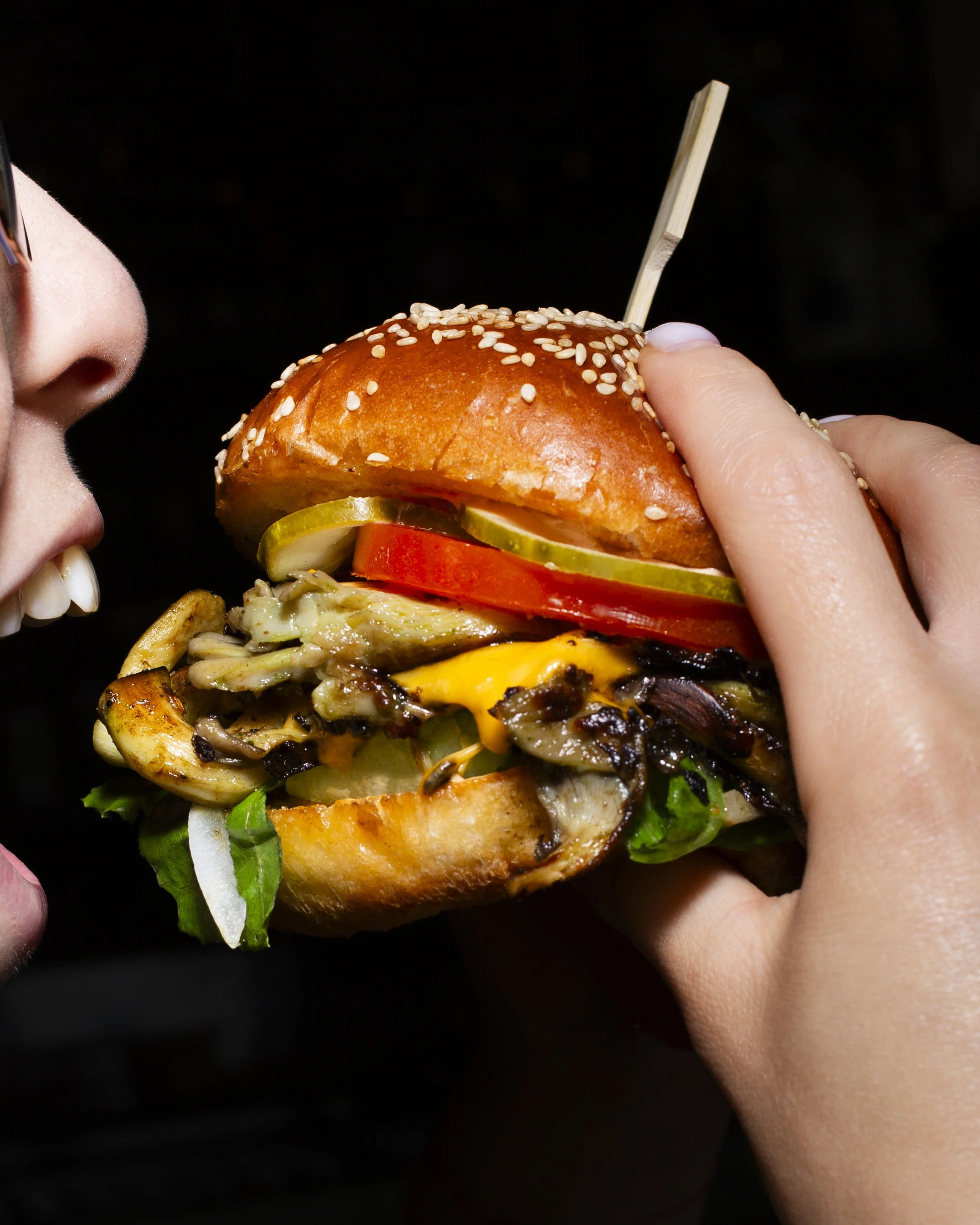 Person about to take a bite of a large gourmet burger with sesame seed bun, pickles, tomato, lettuce, cheese, and grilled mushrooms.