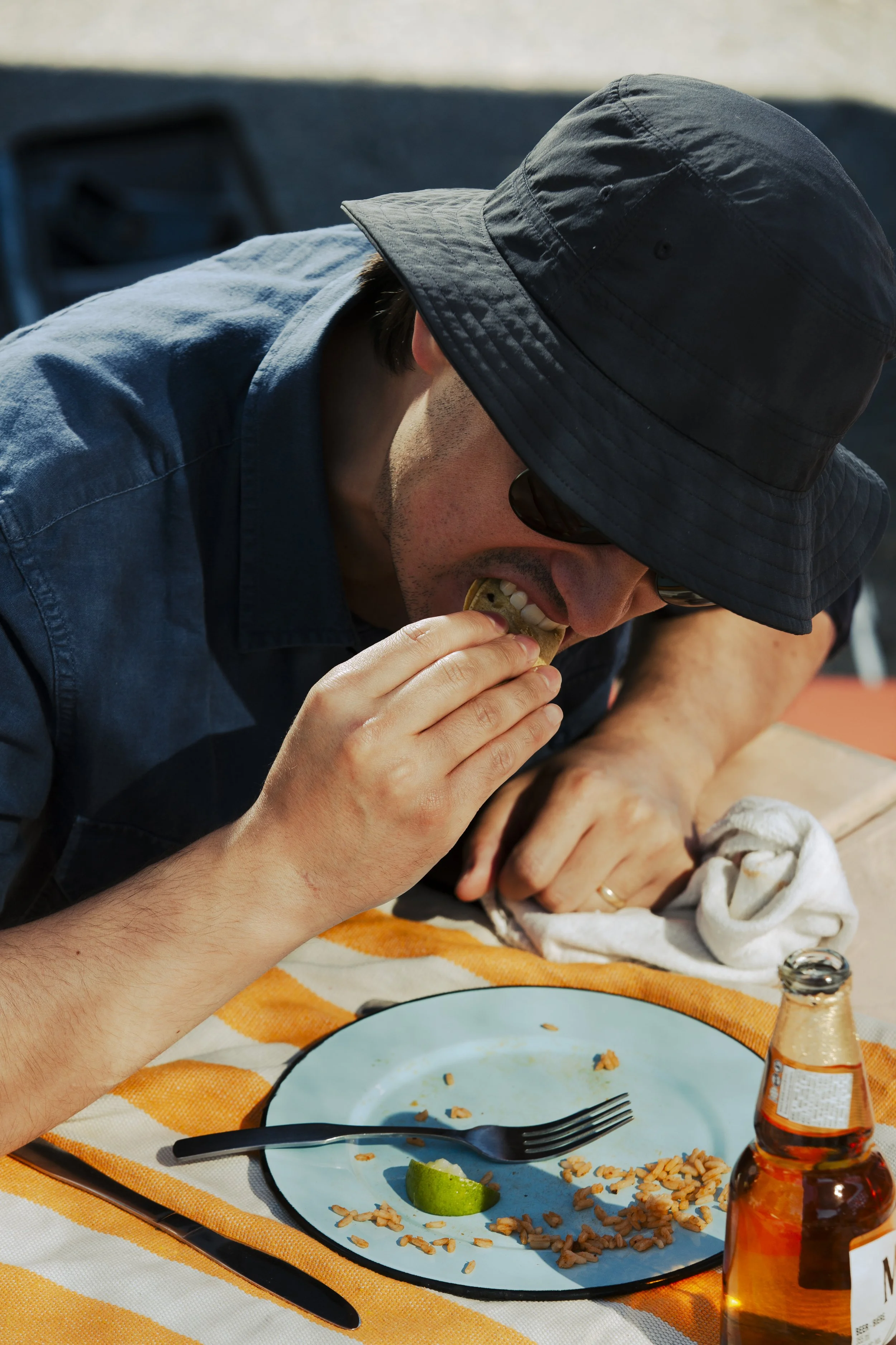 Man wearing a dark bucket hat and sunglasses eating food at an outdoor table with a lime wedge and a beer bottle.
