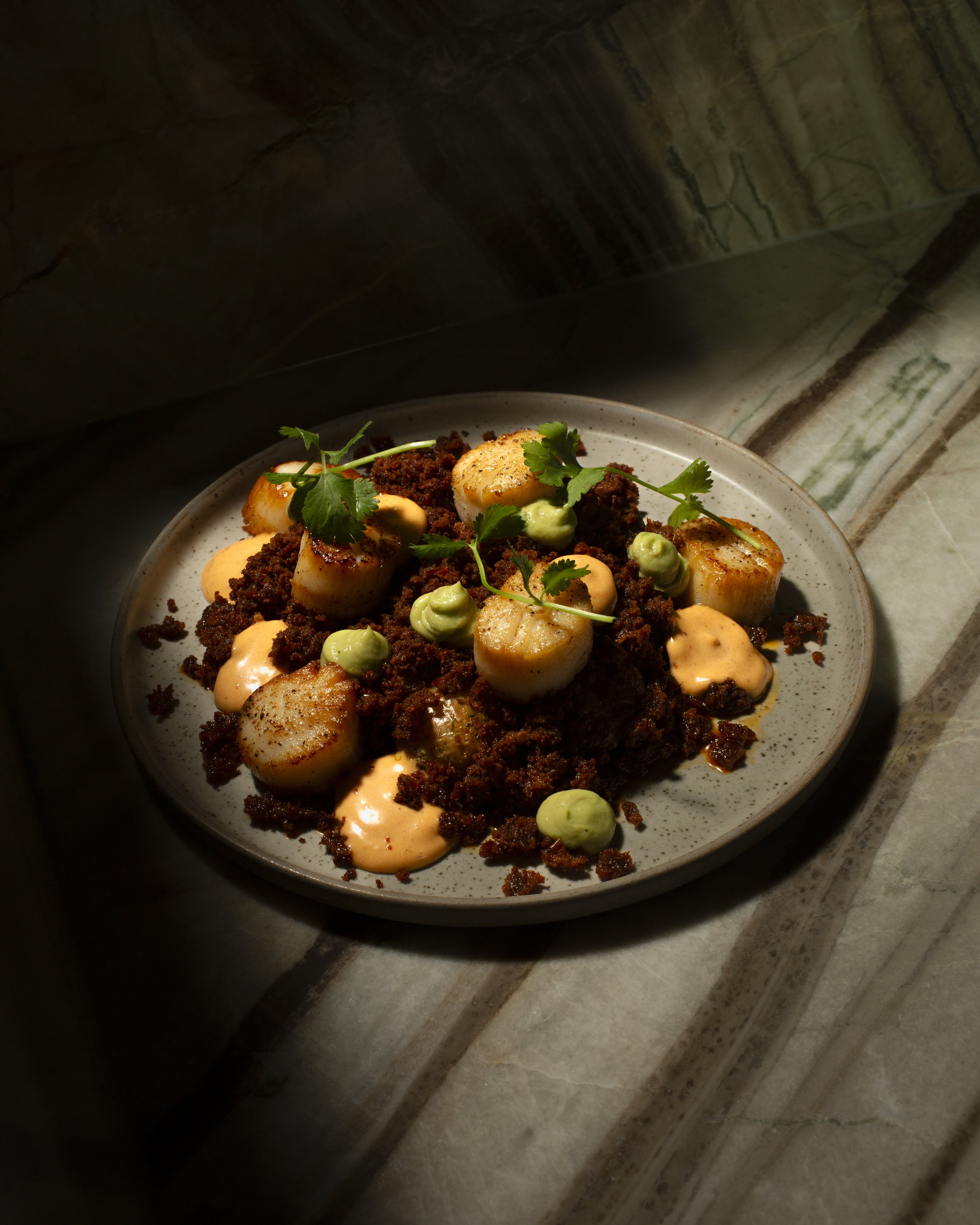 A plate of seared scallops with a bed of ground meat, garnished with cilantro and a drizzle of creamy sauce.