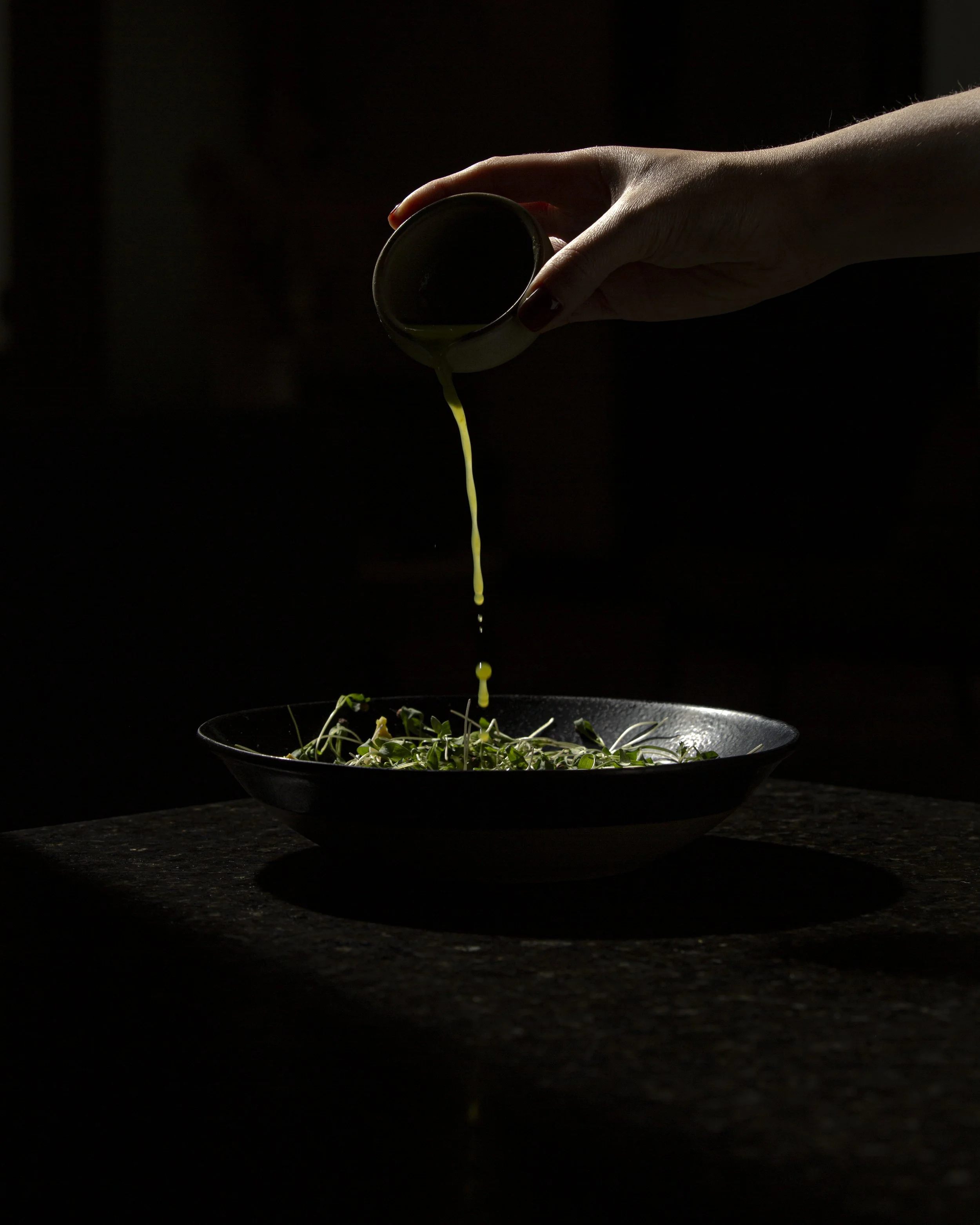 A person pouring olive oil over a salad of greens in a black bowl, with a dark background.