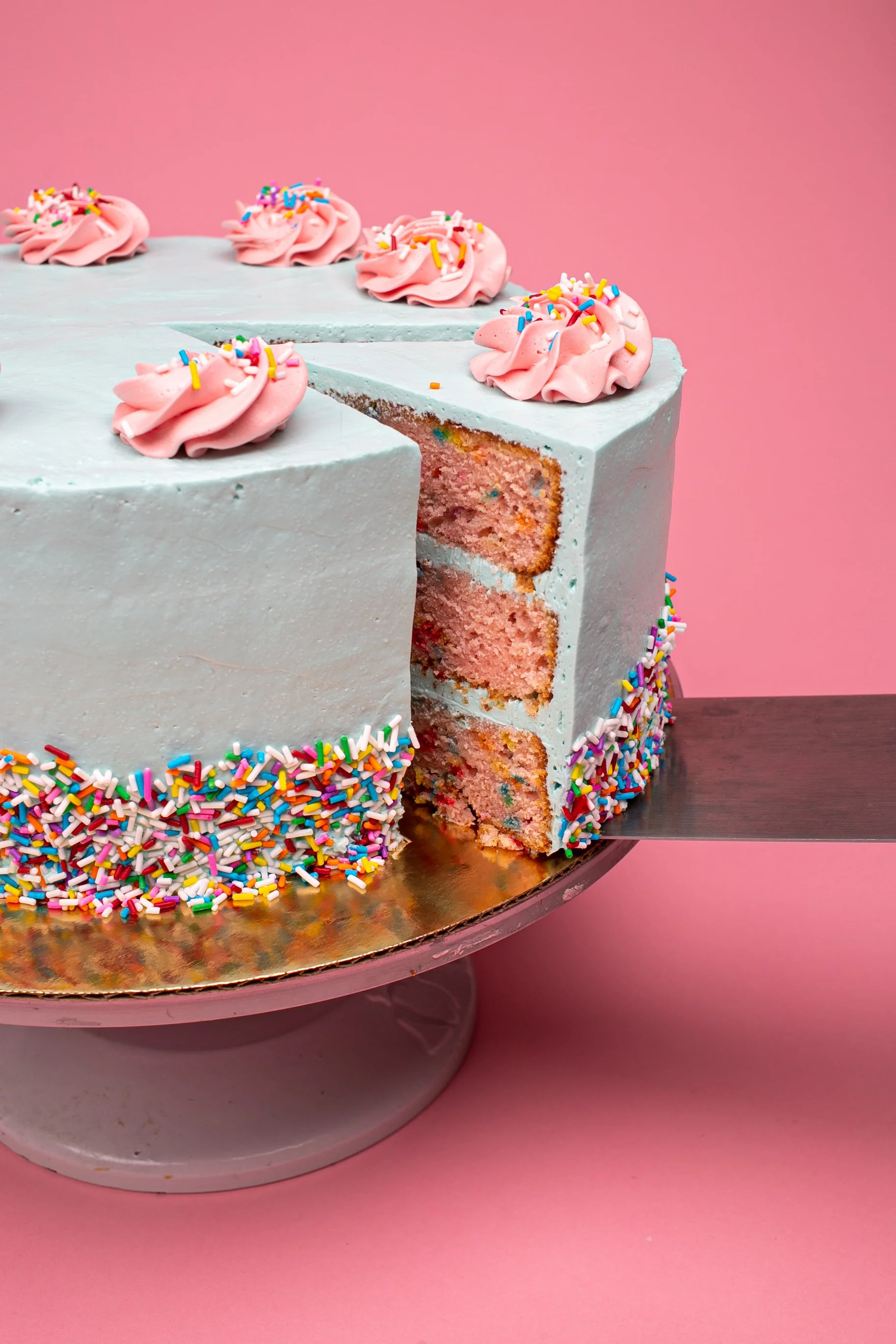 A round birthday cake with white frosting decorated with colorful rainbow sprinkles and pink frosting rosettes with sprinkles on top, with a slice being served and the interior showing pink cake with colorful sprinkles, set against a pink background.