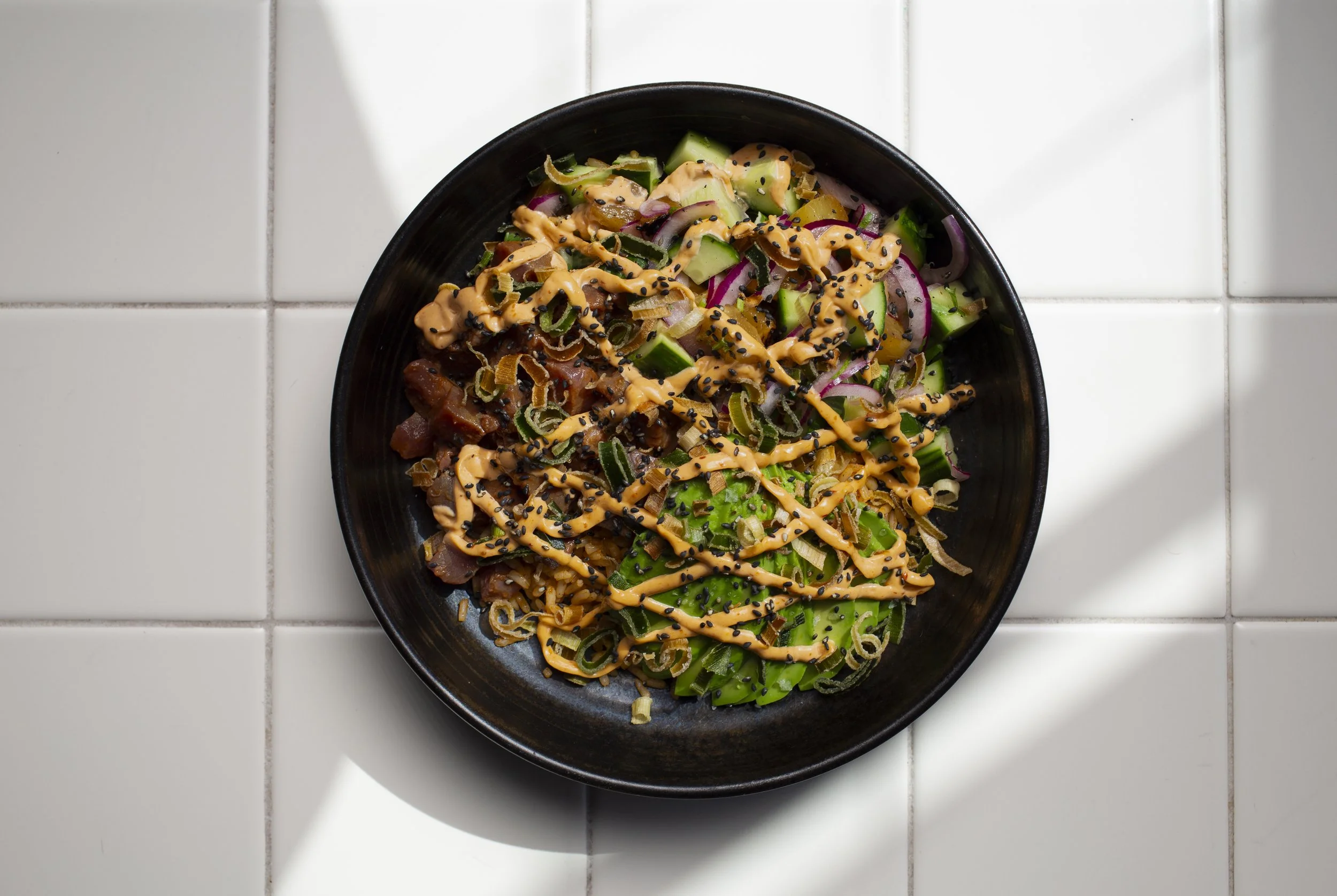 Black bowl of poke salad with sliced cucumbers, red onions, green onions, seaweed, and drizzled spicy mayonnaise on white tiled surface
