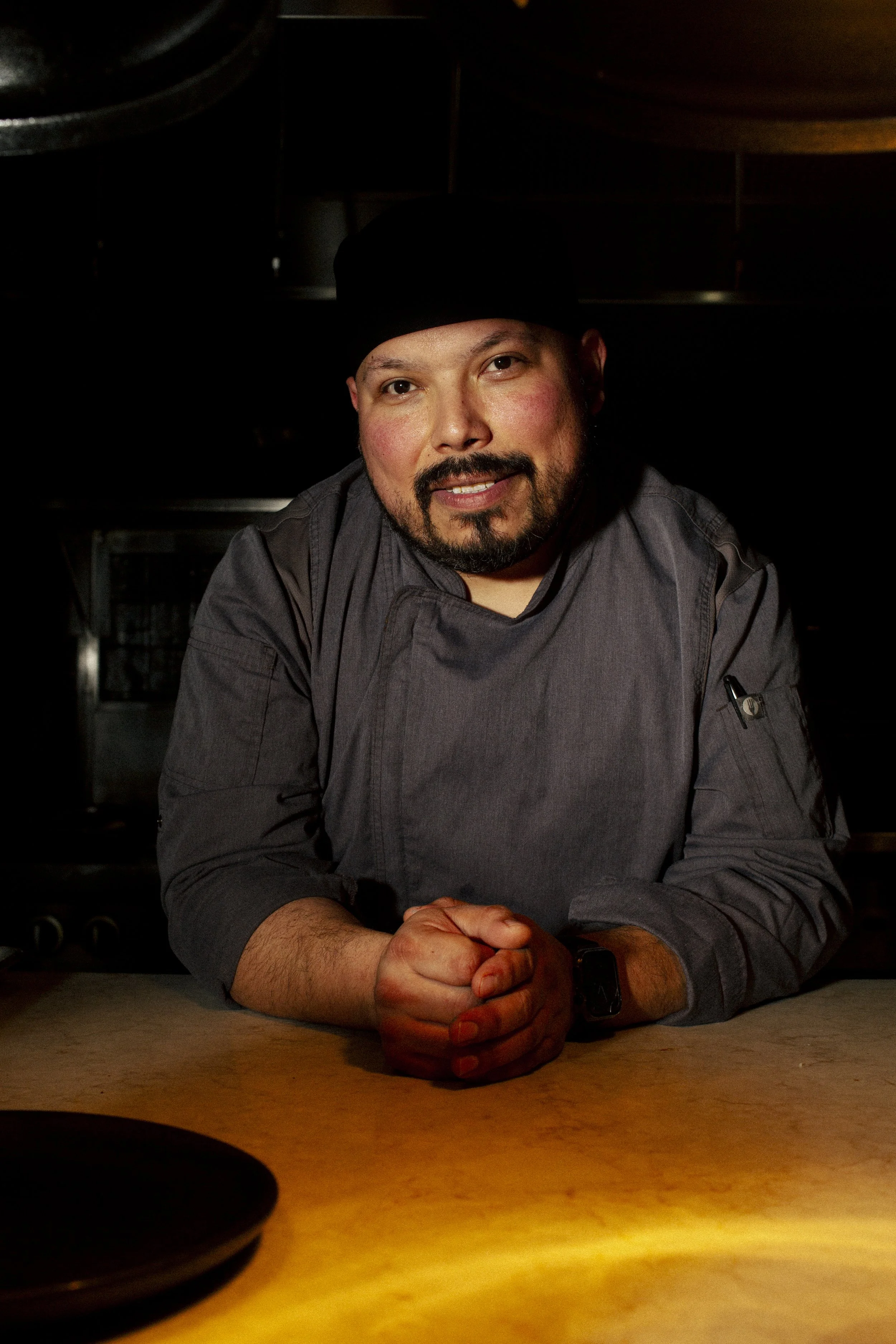 A male chef in a black hat and dark gray chef jacket leaning on a counter with hands clasped, smiling in a dimly lit kitchen.