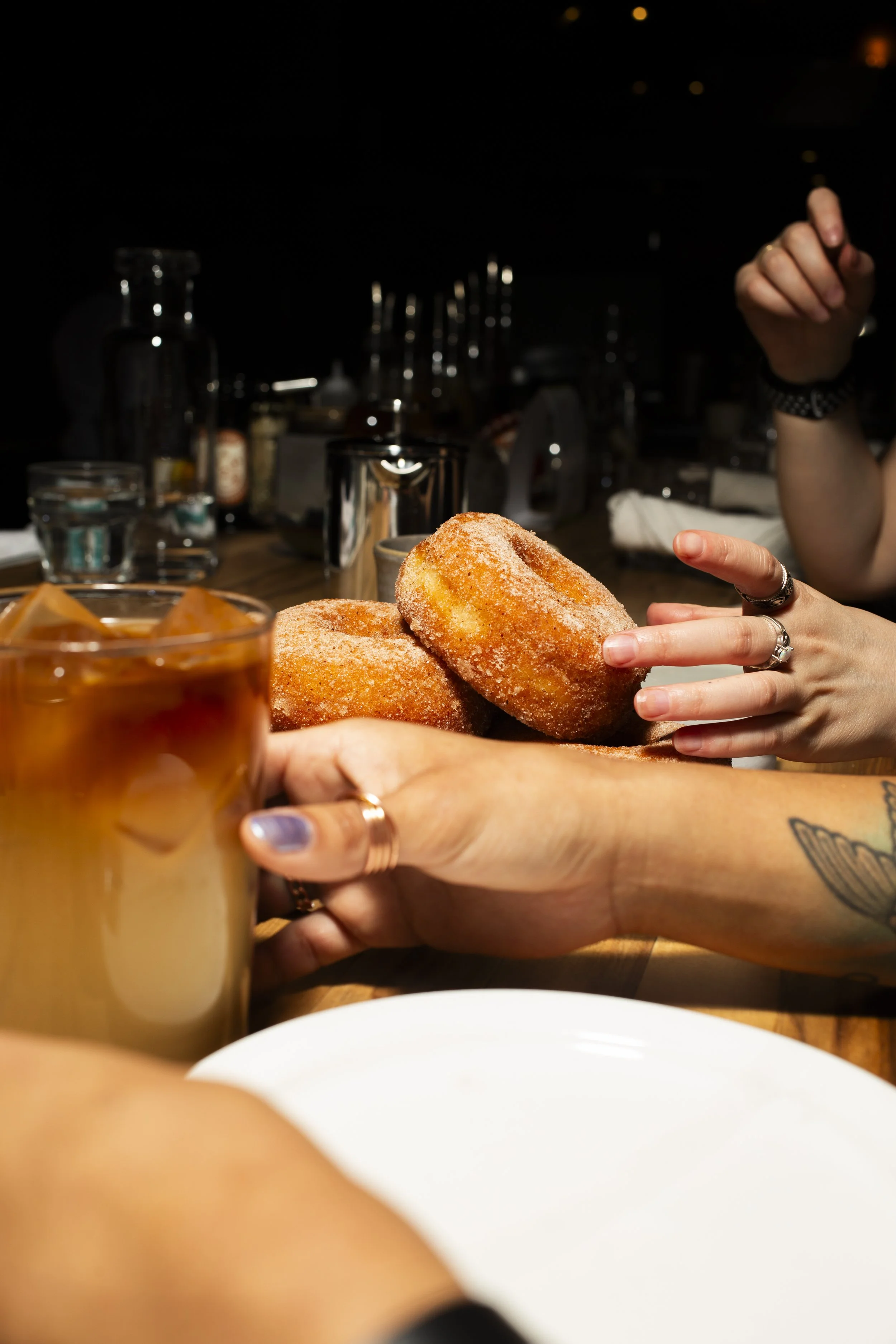 Person holding a glass of iced coffee or tea, reaching for sugar-dusted donuts on a plate, with a dark restaurant or bar background.
