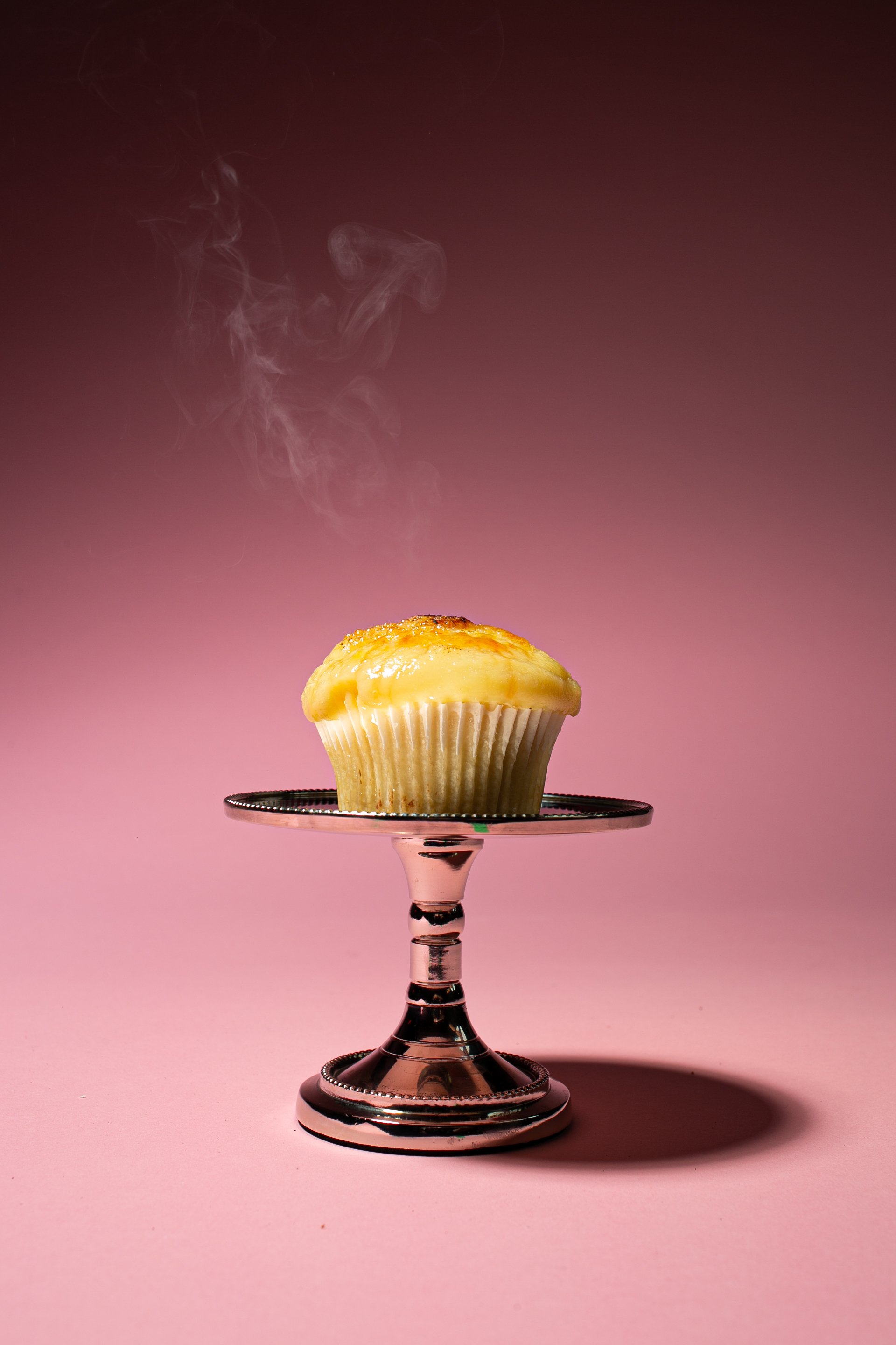 A steaming cupcake on a silver pedestal against a pink background.