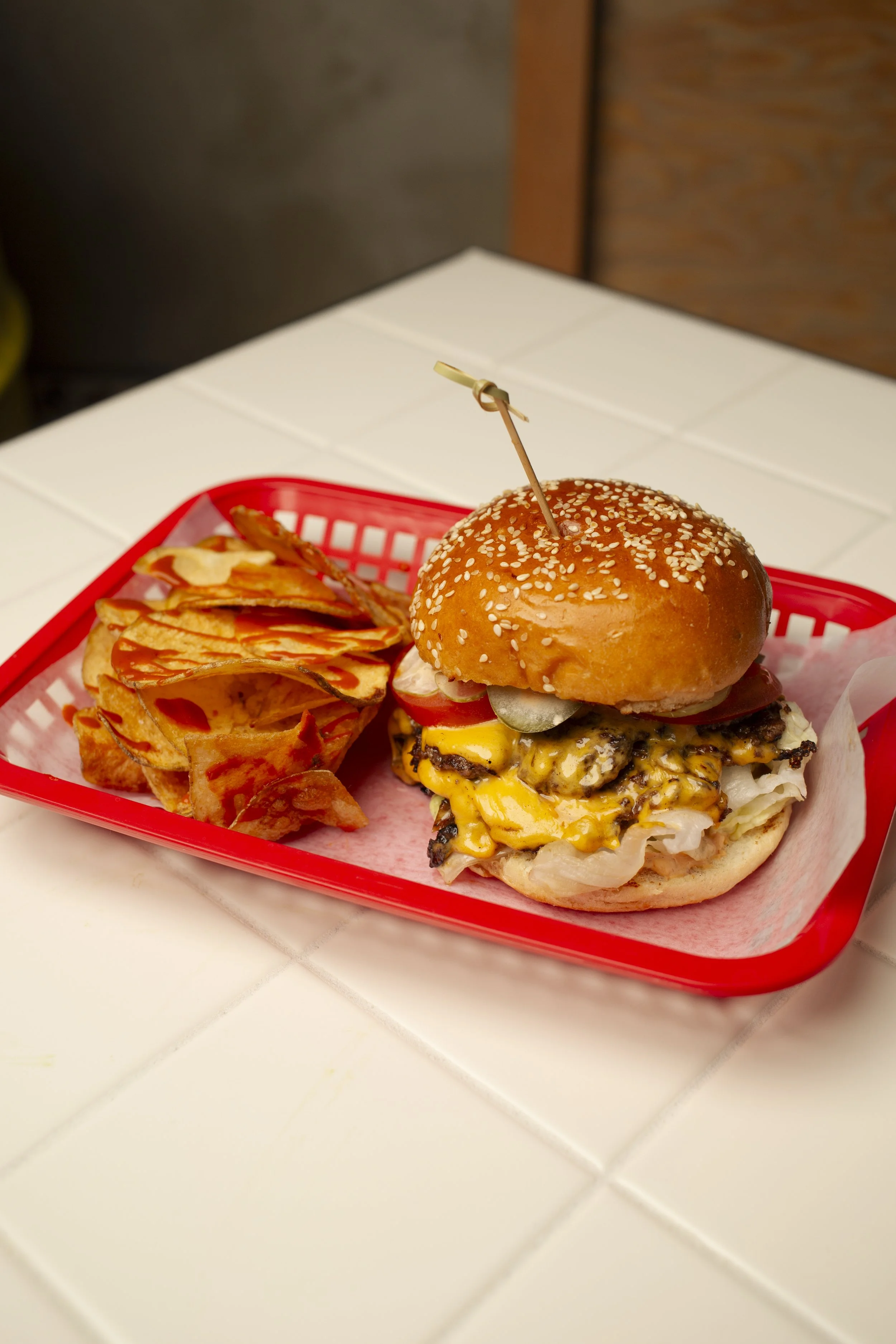 A cheeseburger with lettuce, tomato, pickles, and onions in a sesame seed bun, served with a side of potato chips on a red tray.