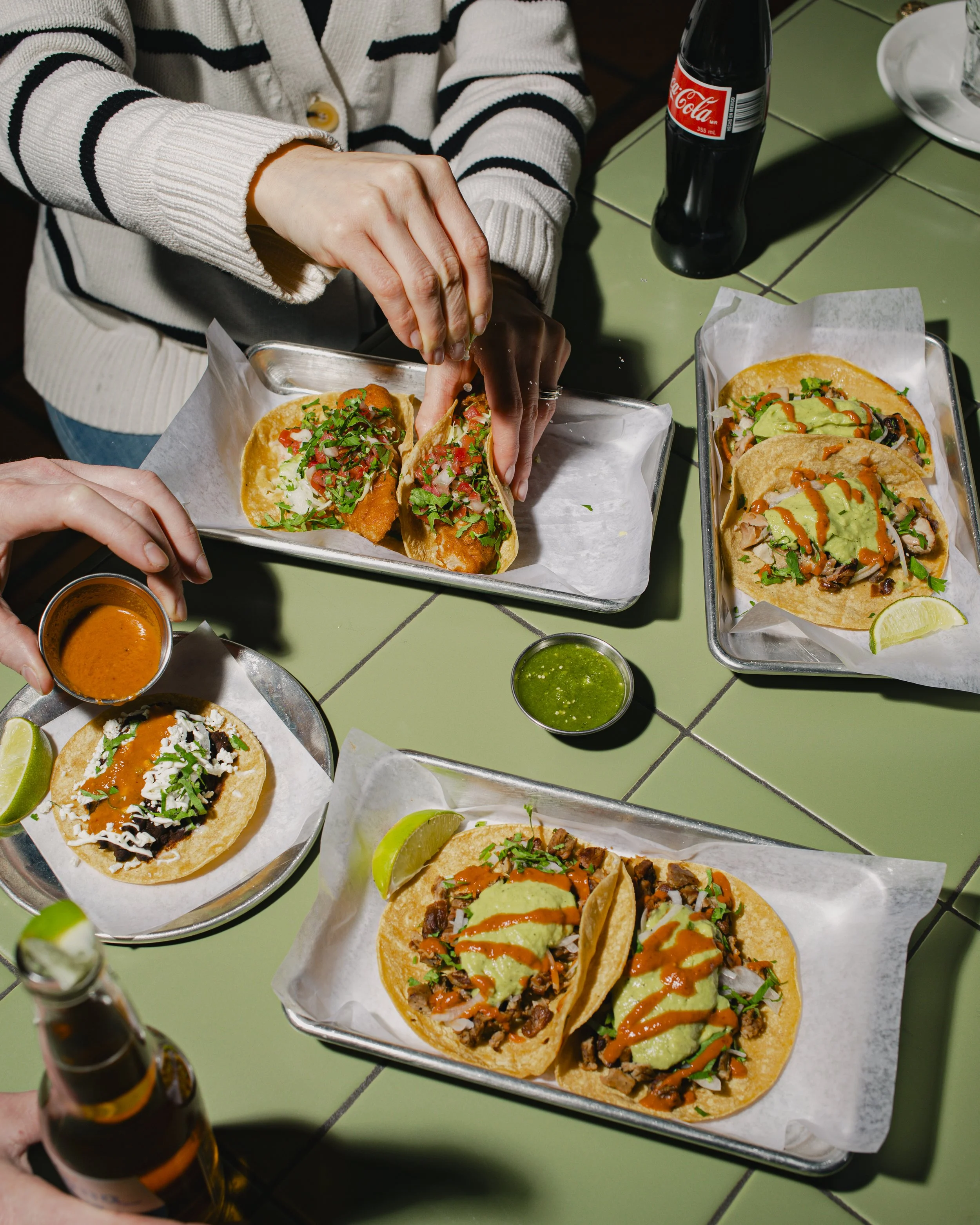 People sharing Mexican tacos and drinks at a restaurant table with green tiled surface, including tacos with various fillings, lime wedges, small bowls of salsa, and bottles of soda.