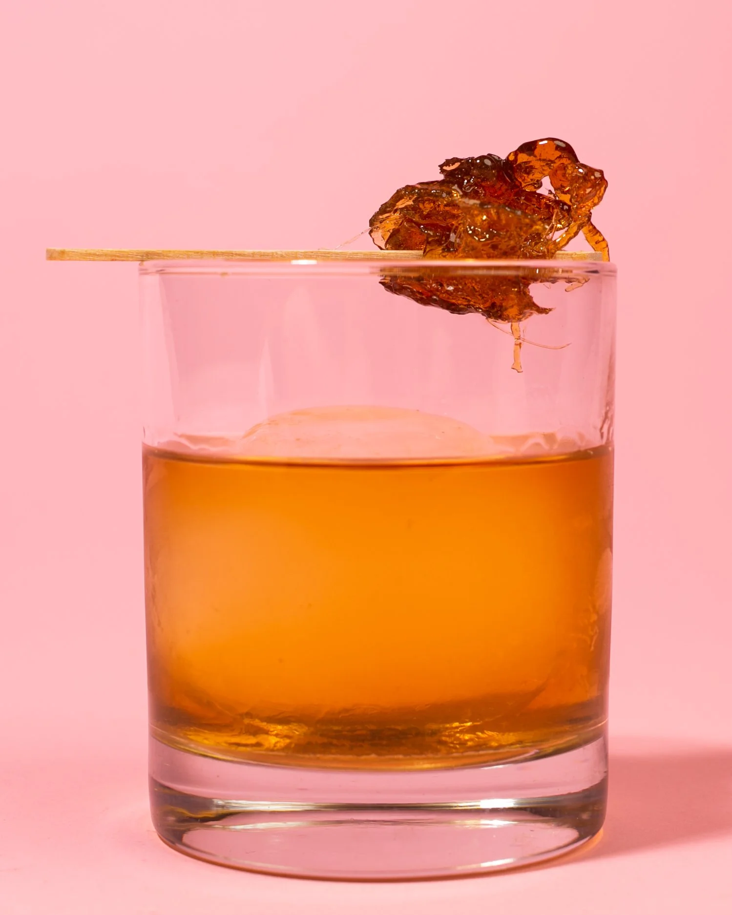 A glass of amber-colored cocktail with a honey-colored sugar cube on a toothpick balanced on the rim, against a pink background.