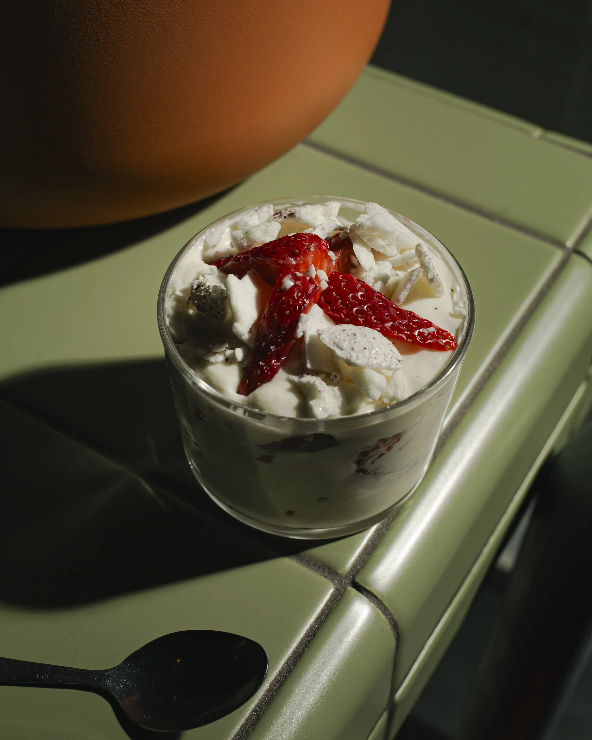 A glass of strawberry shortcake with whipped cream and sliced strawberries on top, placed on a green-tiled surface with a terracotta pot in the background.