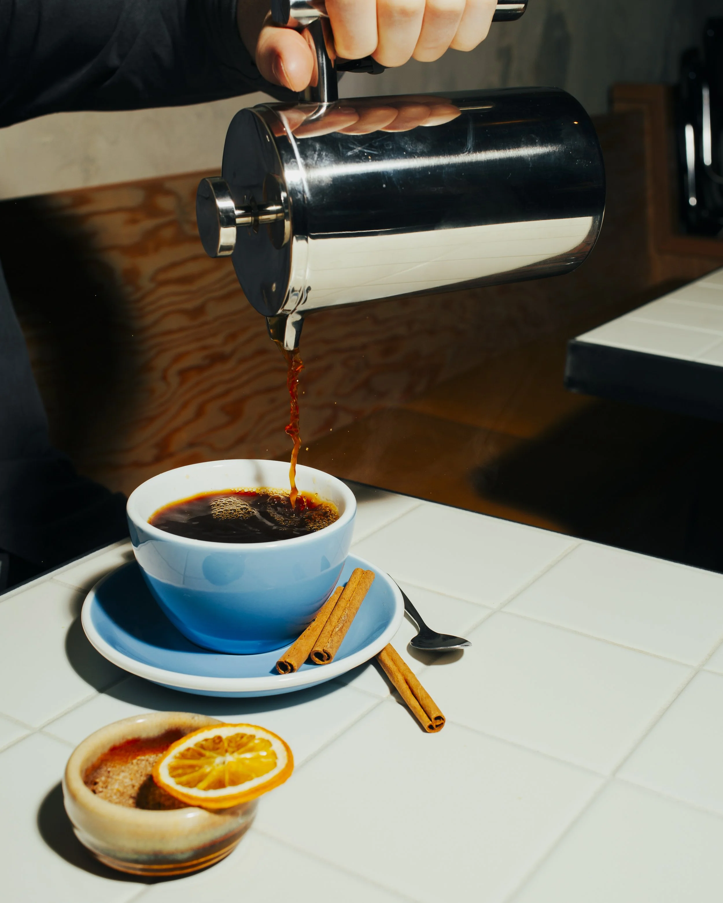 A person is pouring coffee from a metallic coffee pot into a white coffee cup on a saucer with two cinnamon sticks. There is also a small bowl with a slice of orange on the tiled counter.