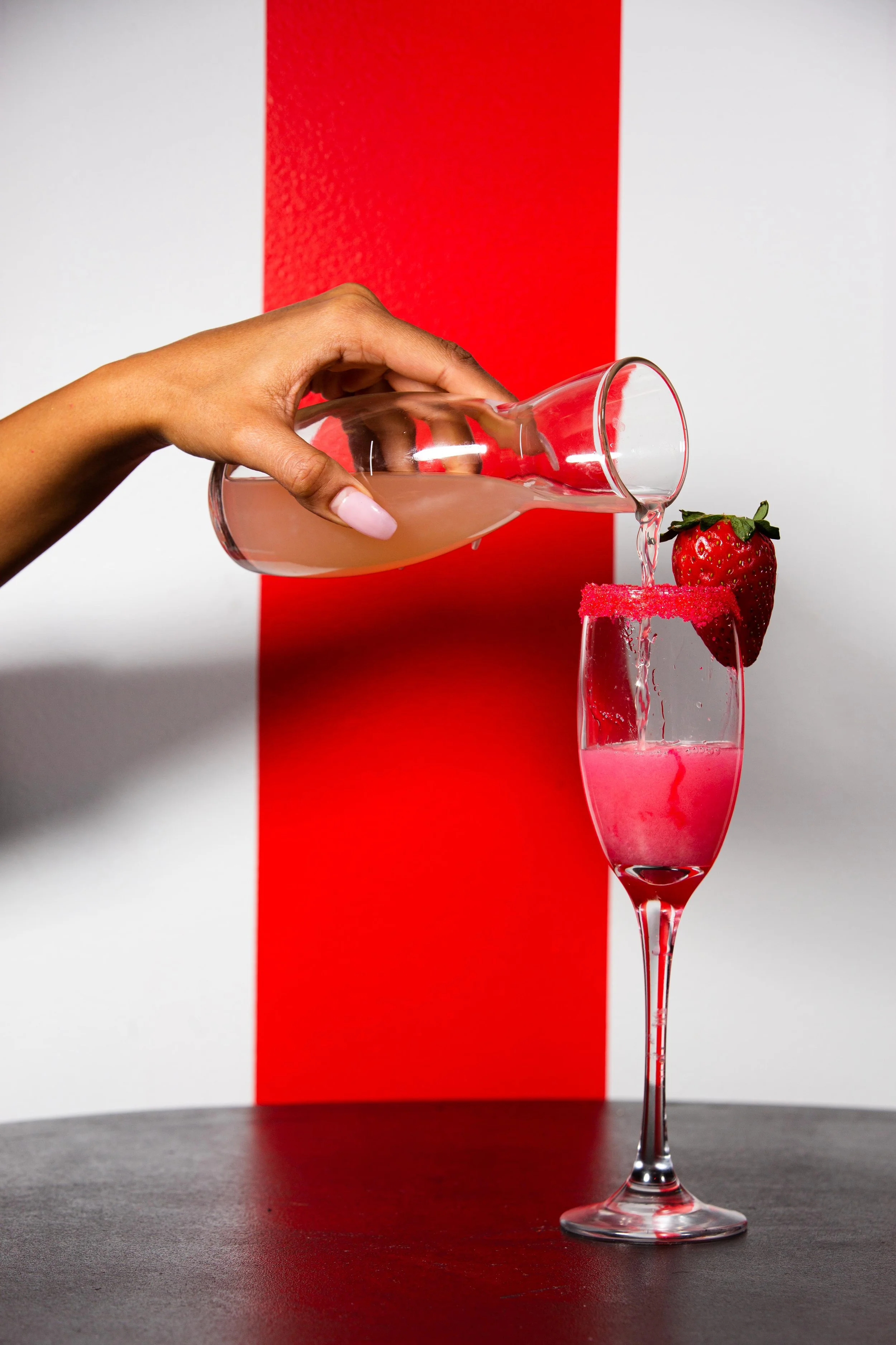 A hand pouring pink cocktail from a glass pitcher into a tall glass rimmed with pink sugar, with a strawberry garnish, against a red and white background.
