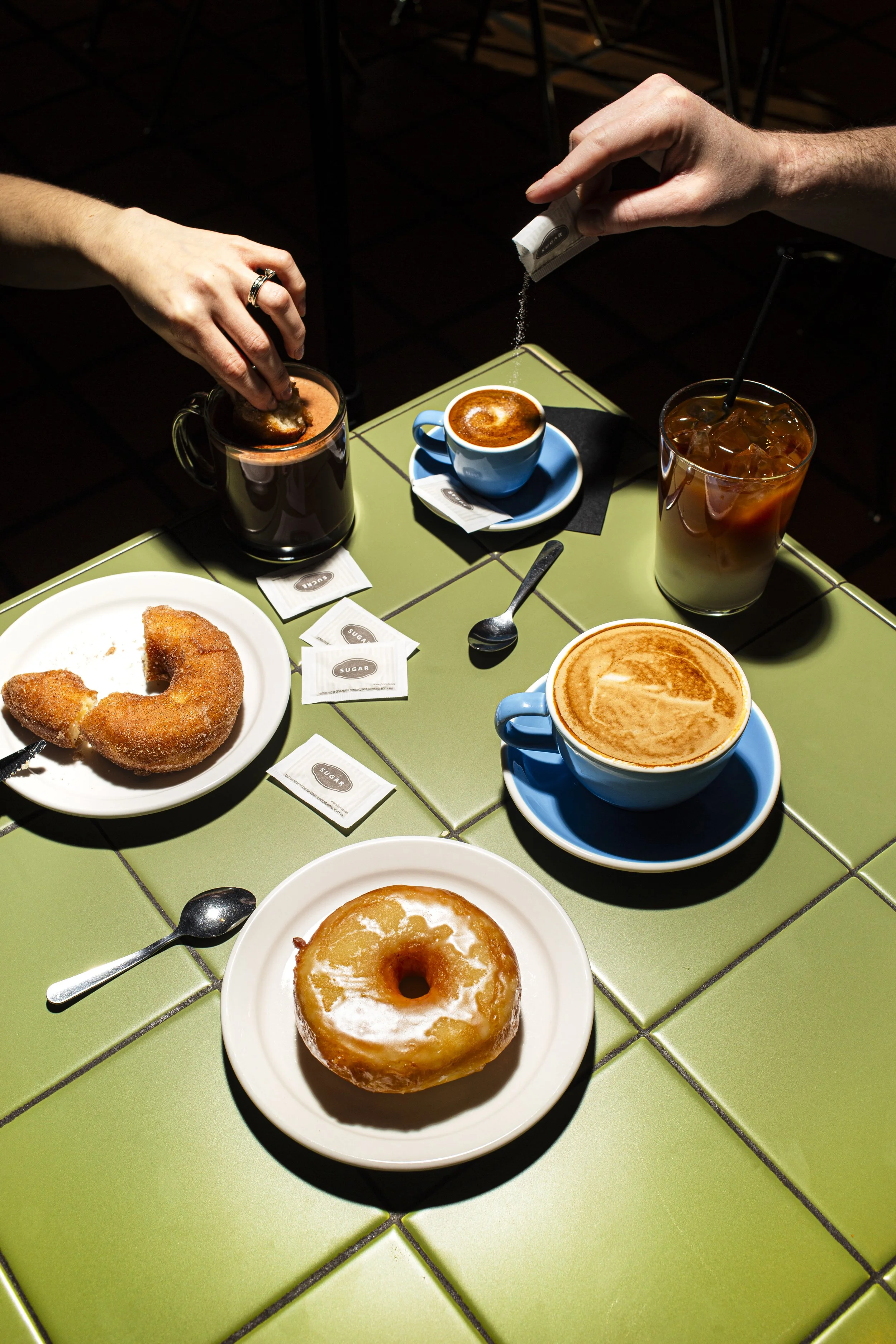 A table with two cups of coffee, a glass of iced coffee, a plate with a donut, a sugar packet, and two hands, one adding sugar to a coffee.