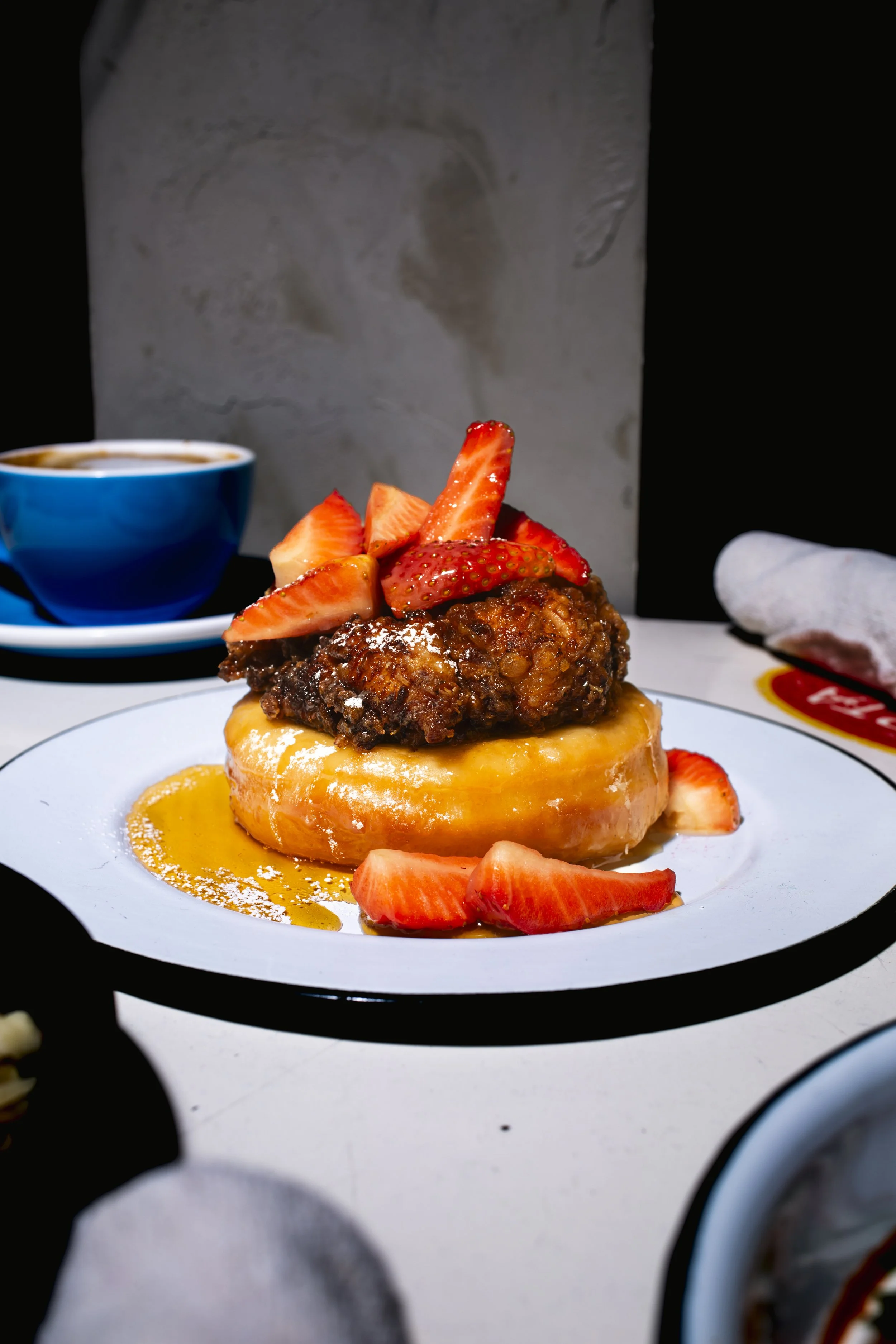 A plate with a doughnut topped with strawberries, fried chicken, powdered sugar, and syrup on a white plate.