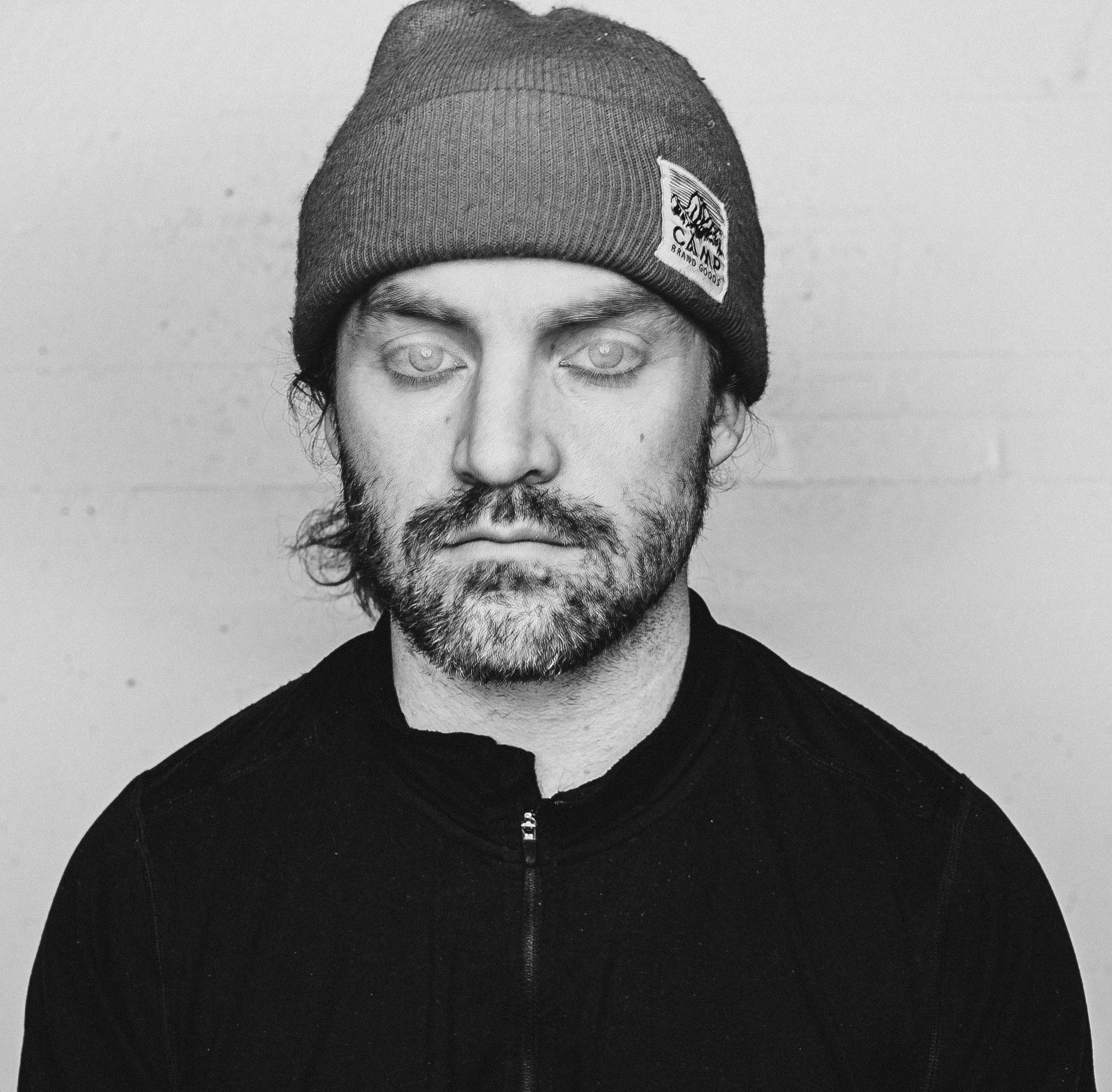 Black and white portrait of a man with a beard wearing a beanie hat and a zip-up jacket, facing forward with a serious expression.