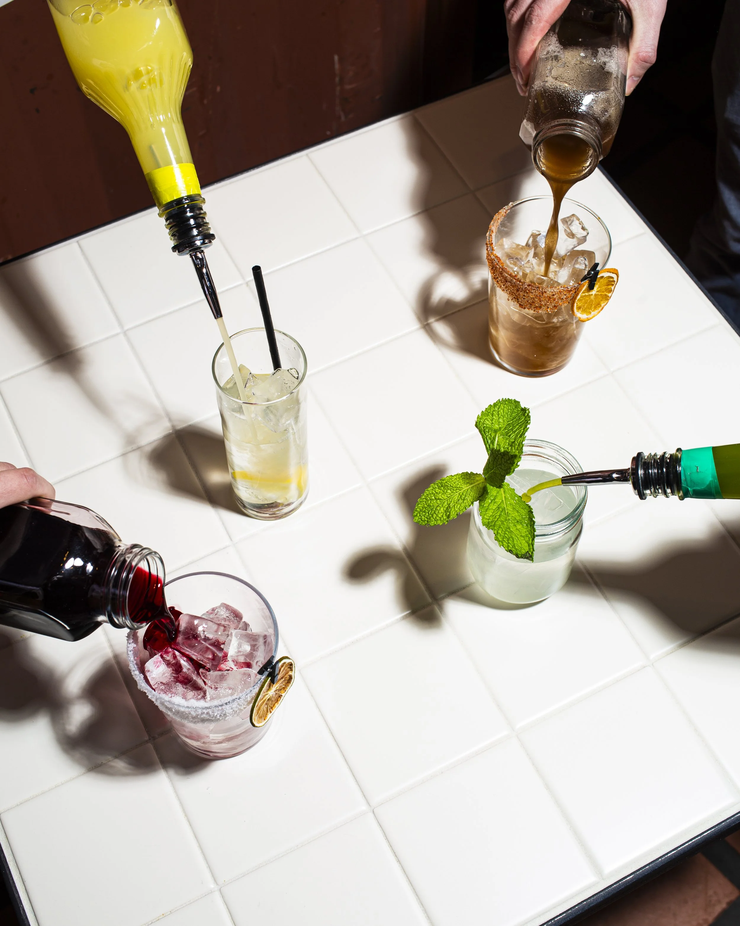 Four colorful cocktails with various garnishes on a white tiled table, with bartenders pouring liquid into glasses.
