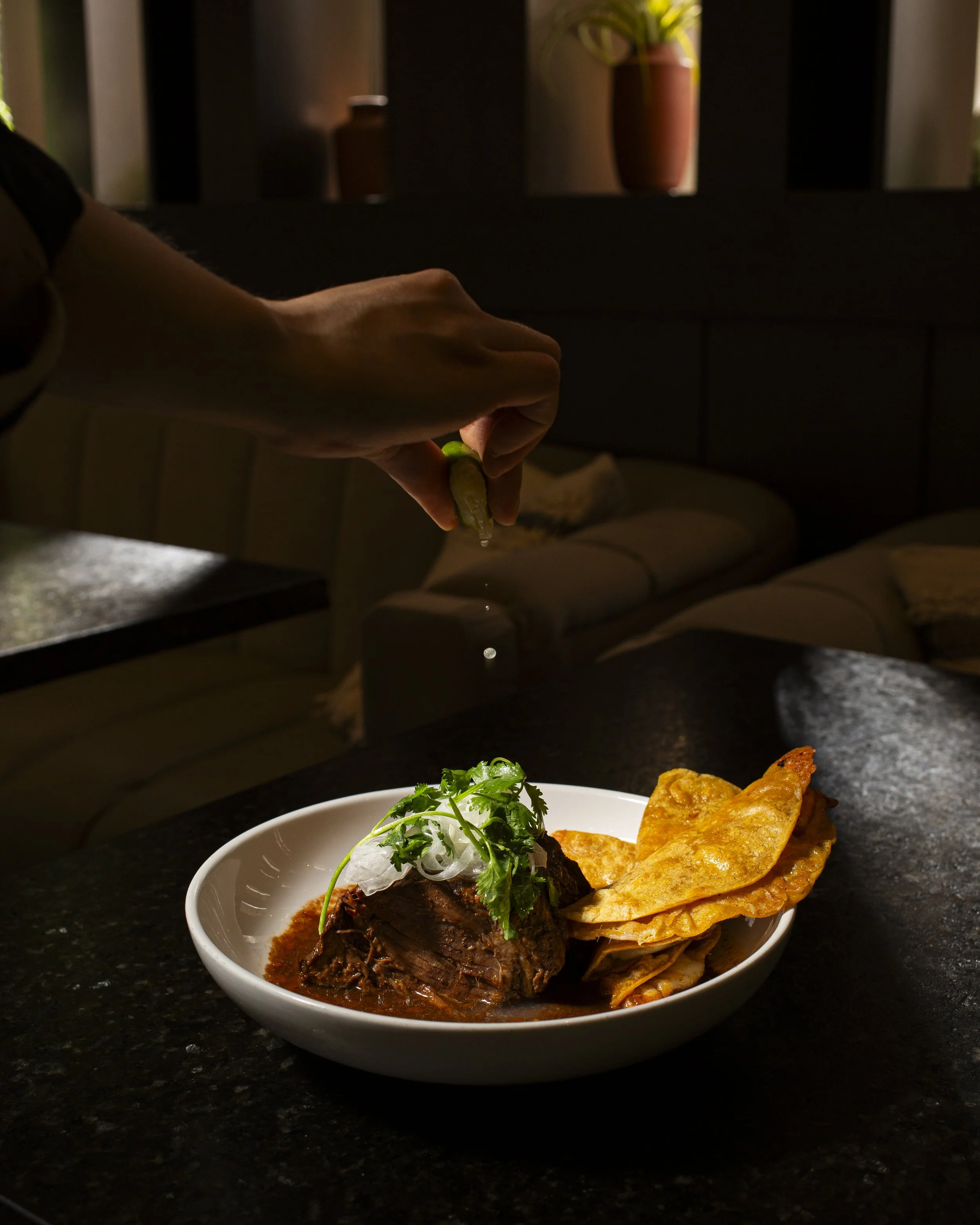 Person sprinkling salt onto a plate of beef stew topped with cilantro and onion, served with fried flatbread on the side, on a dark countertop in a dimly lit room.