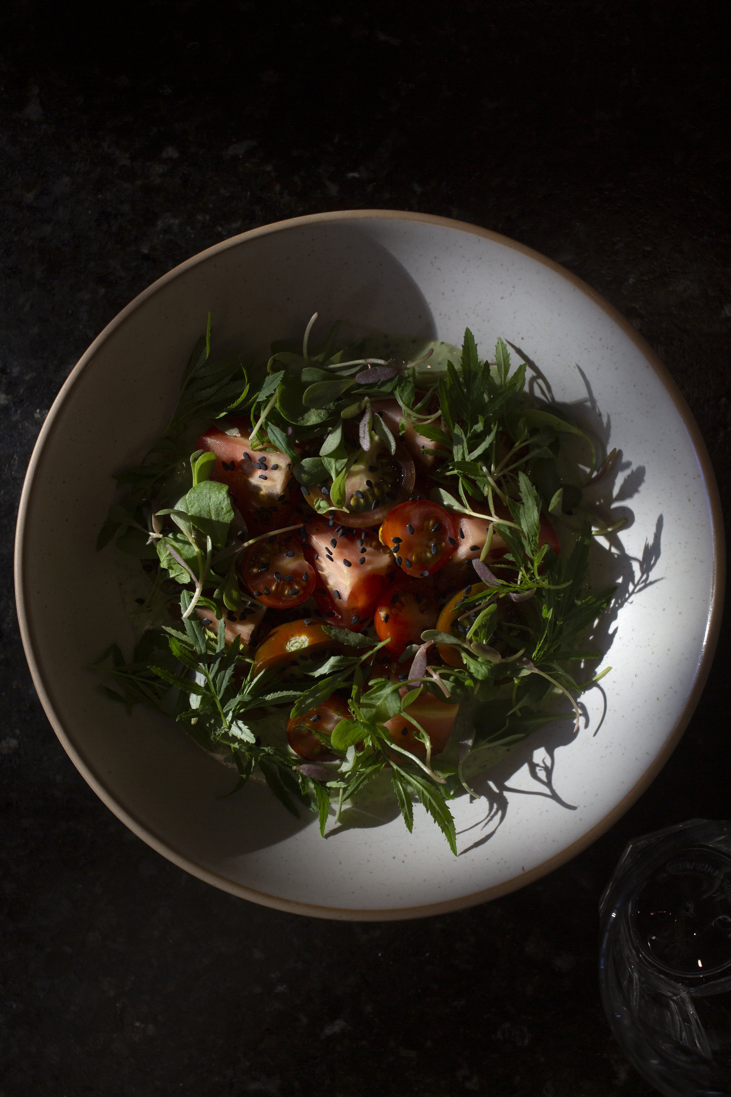 Salad with cherry tomatoes, greens, and seeds in a white bowl on a dark surface.