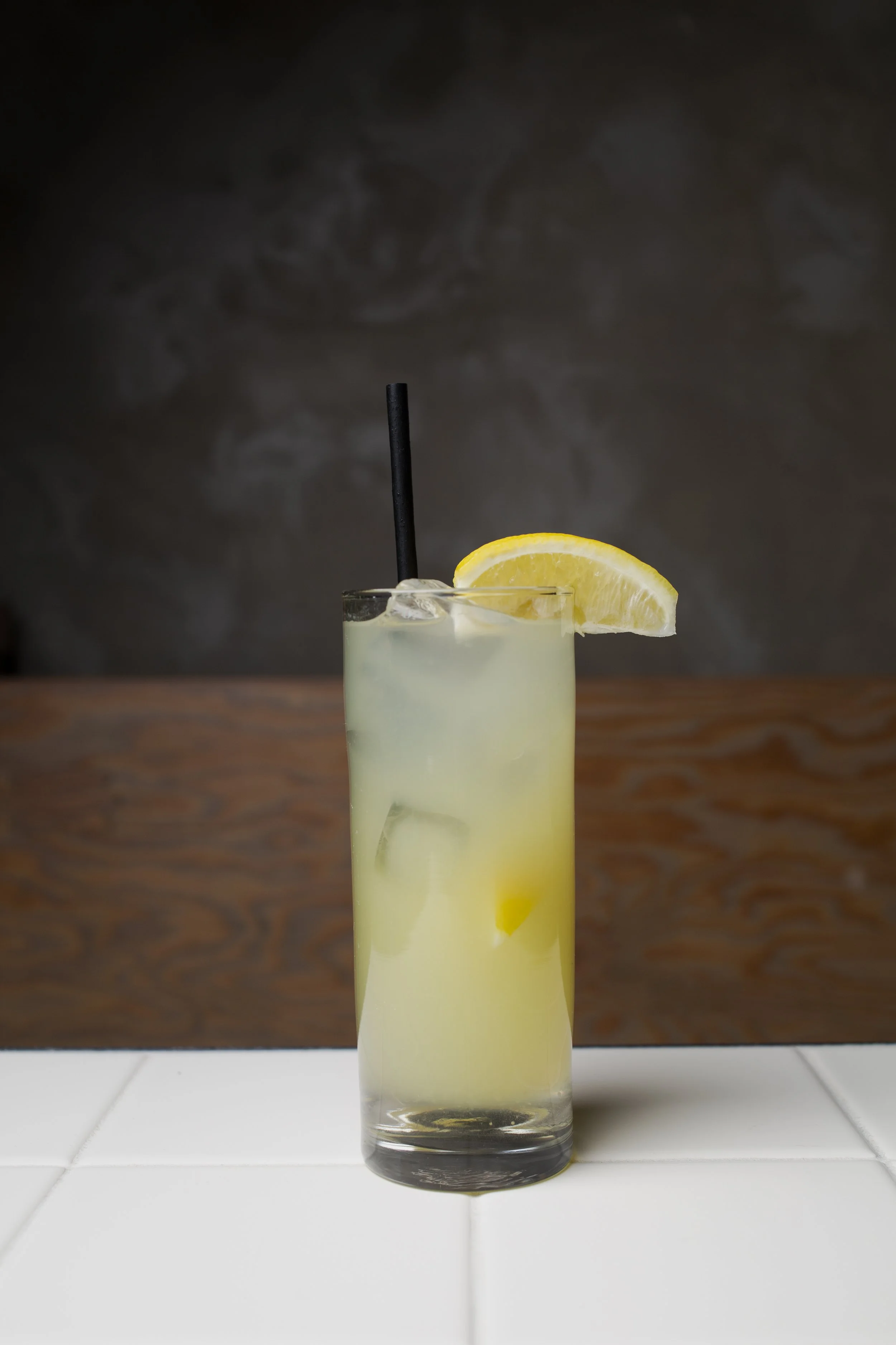 A tall glass of lemonade with ice, garnished with a lemon wedge, a black straw, on a white tiled surface against a dark background.