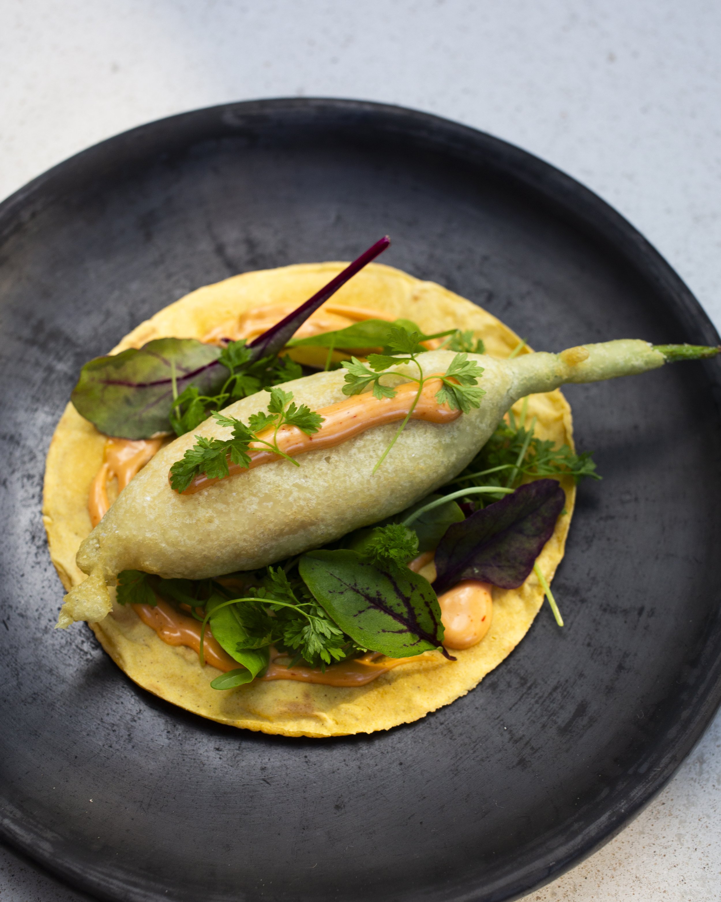 A black plate with a golden-brown fried fish fillet on a bed of colorful mixed greens and topped with a drizzle of orange sauce, garnished with fresh herbs.