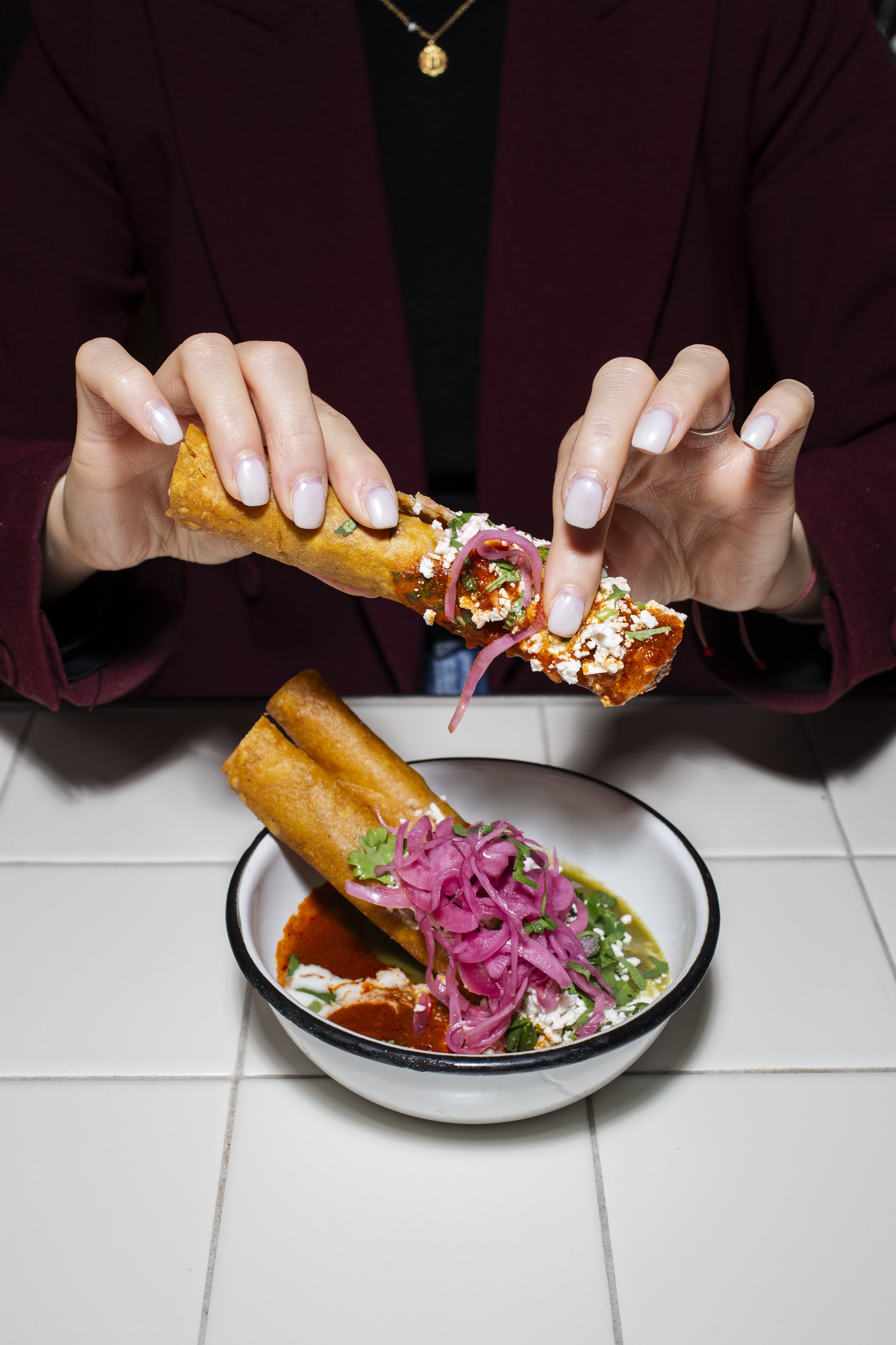 Person holding a tamale over a bowl of similar tamale with red sauce, shredded cheese, cilantro, and pickled red onions.