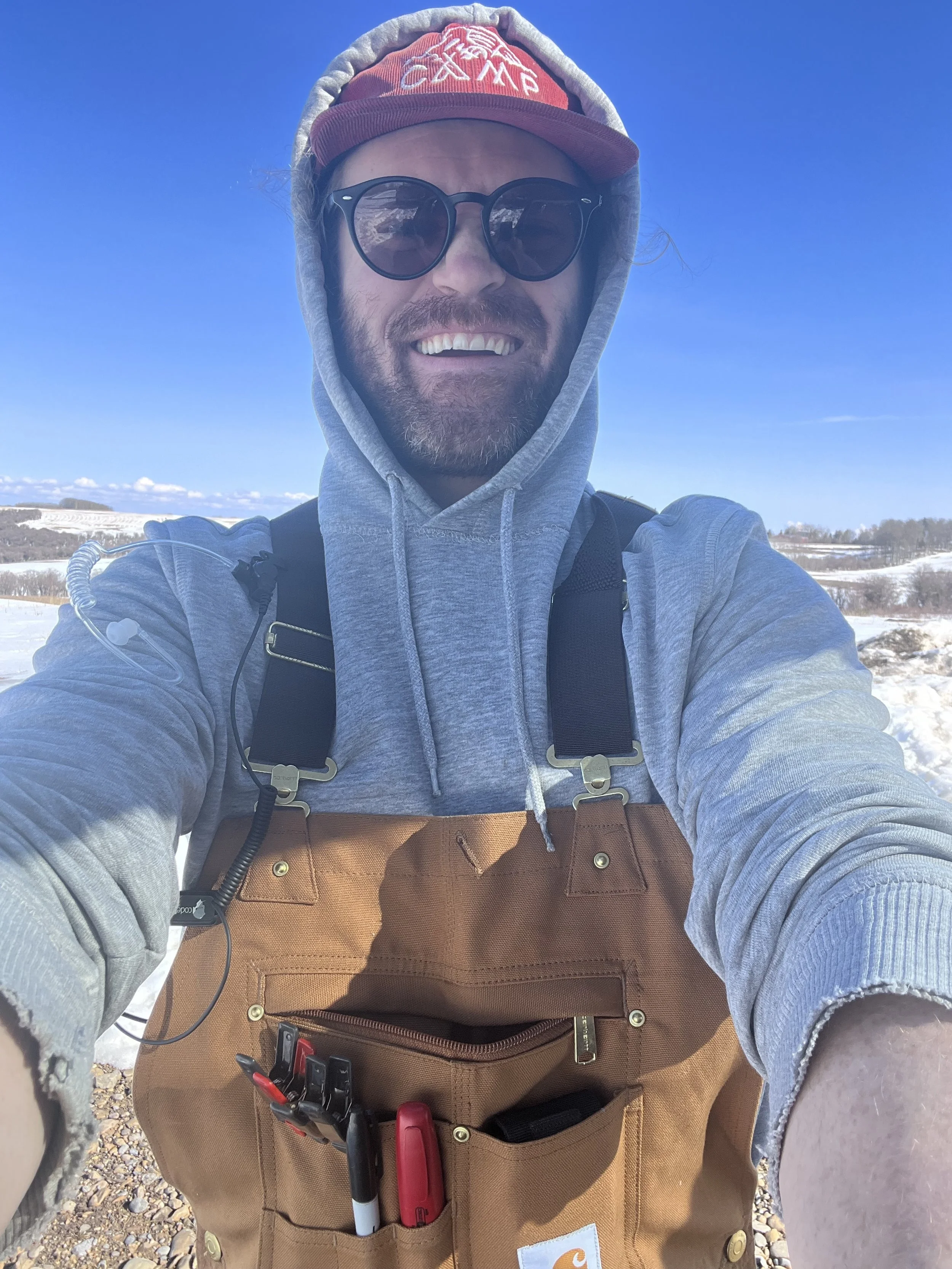 A man outdoors on a snowy landscape, wearing a red baseball cap with white emblem, sunglasses, a gray hoodie, and a brown Carhartt work apron with pens in the pocket, smiling at the camera.