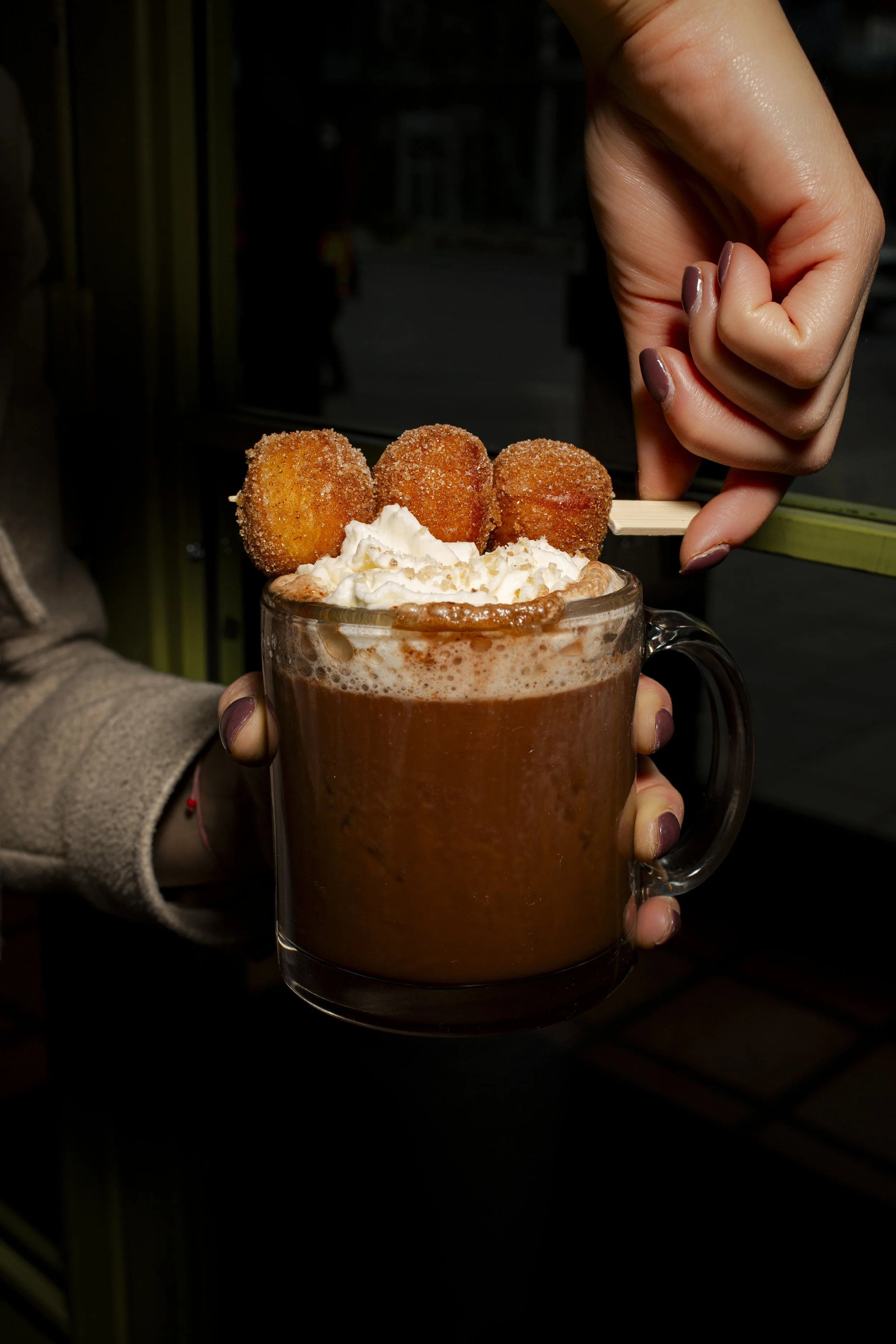 A clear glass mug of hot chocolate topped with whipped cream and three fried dough balls on a skewer, with a person's hand holding the mug.