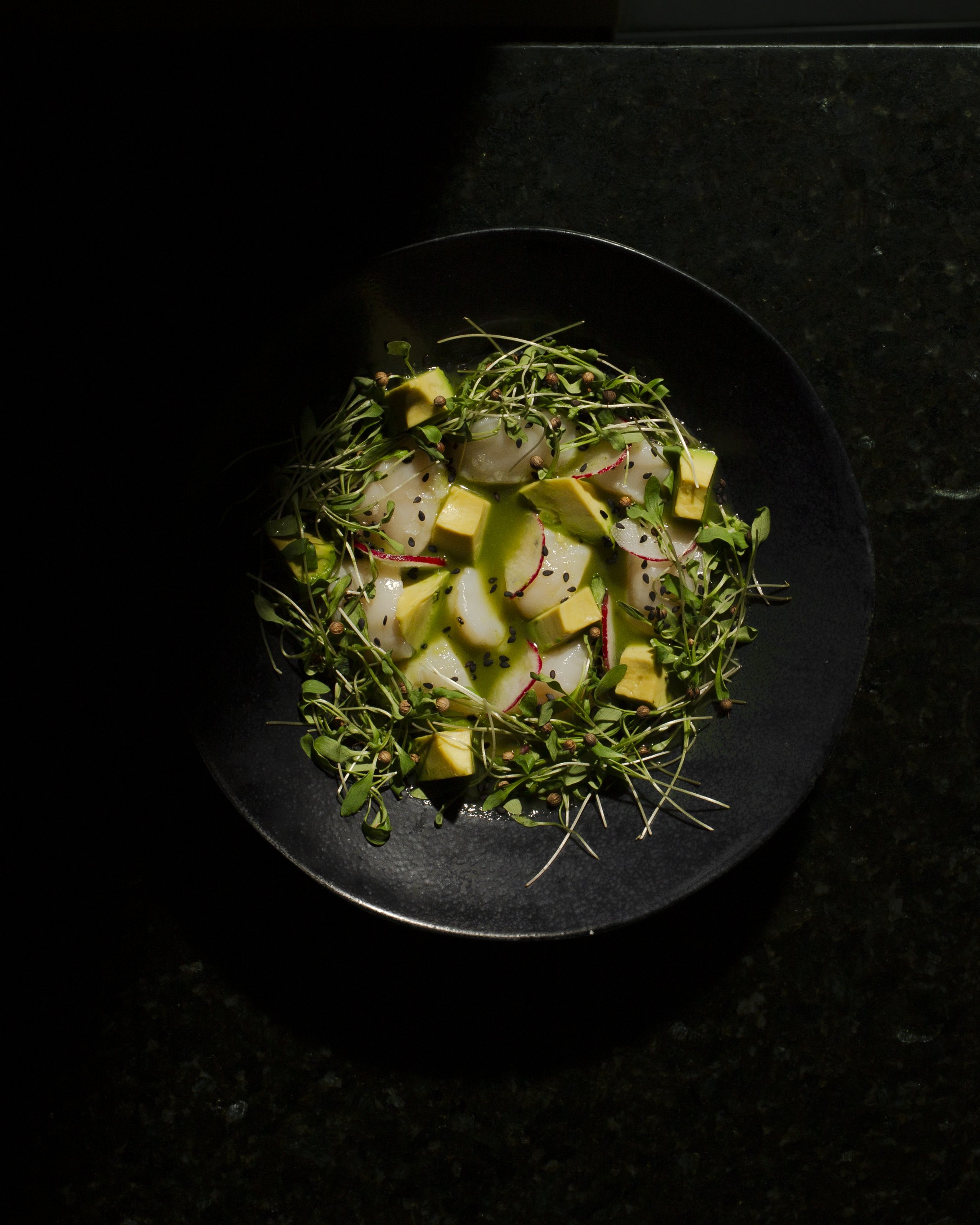 A black bowl of fresh sliced avocado, radish, microgreens, and black sesame seeds on a dark surface.