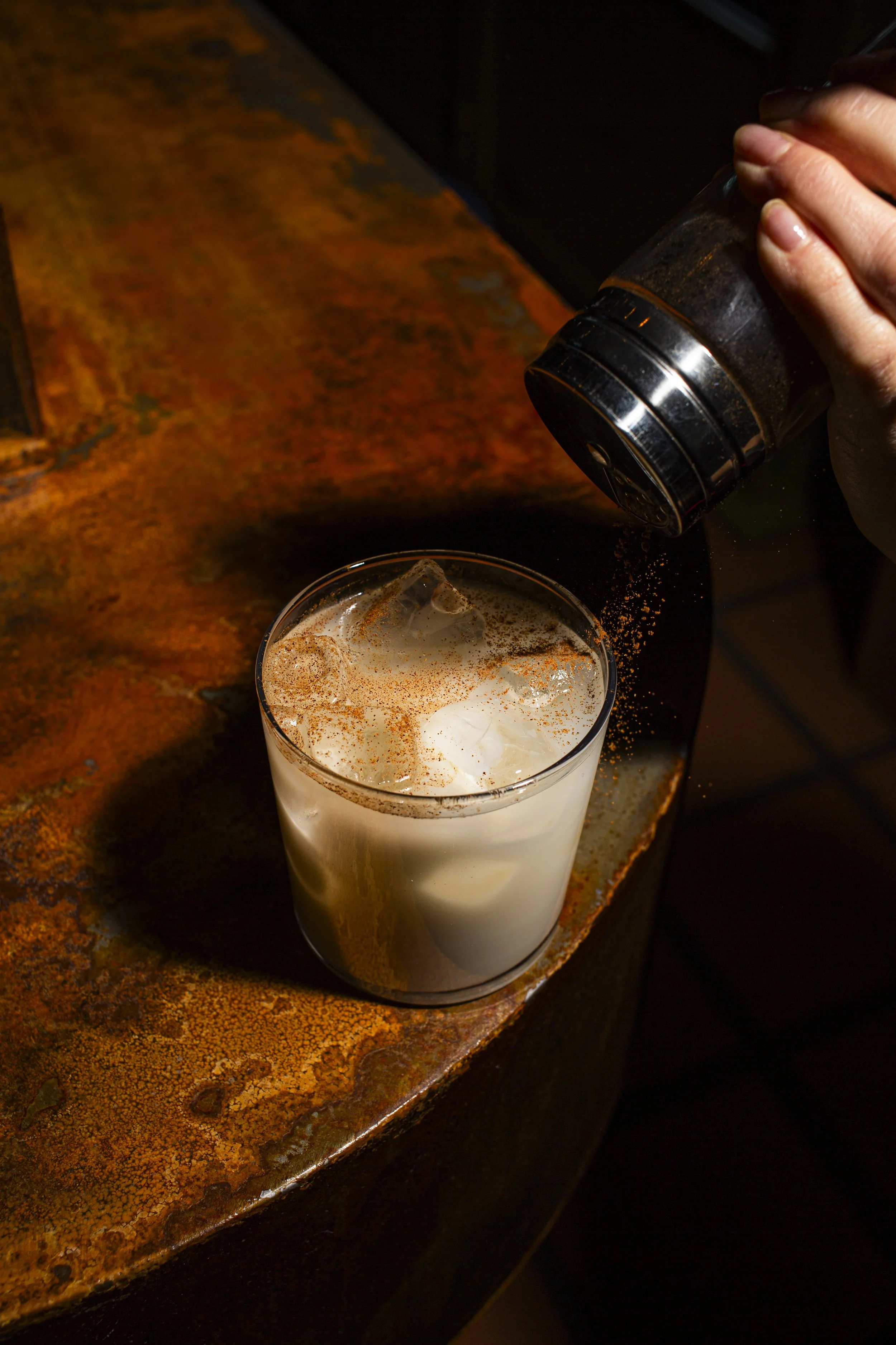 A person sprinkles cinnamon onto a glass of iced horchata, a traditional Mexican beverage, on a rustic wooden surface.