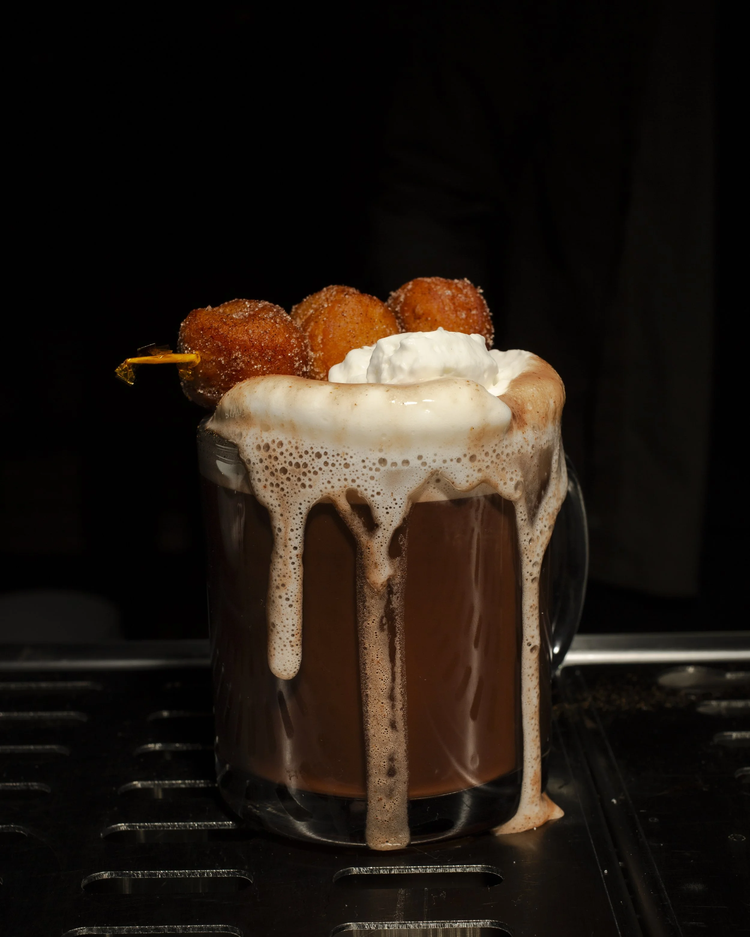 A glass mug filled with a chocolate beverage topped with whipped cream, three deep-fried dessert balls on a skewer, and frothy foam overflowing the mug's rim, against a dark background.