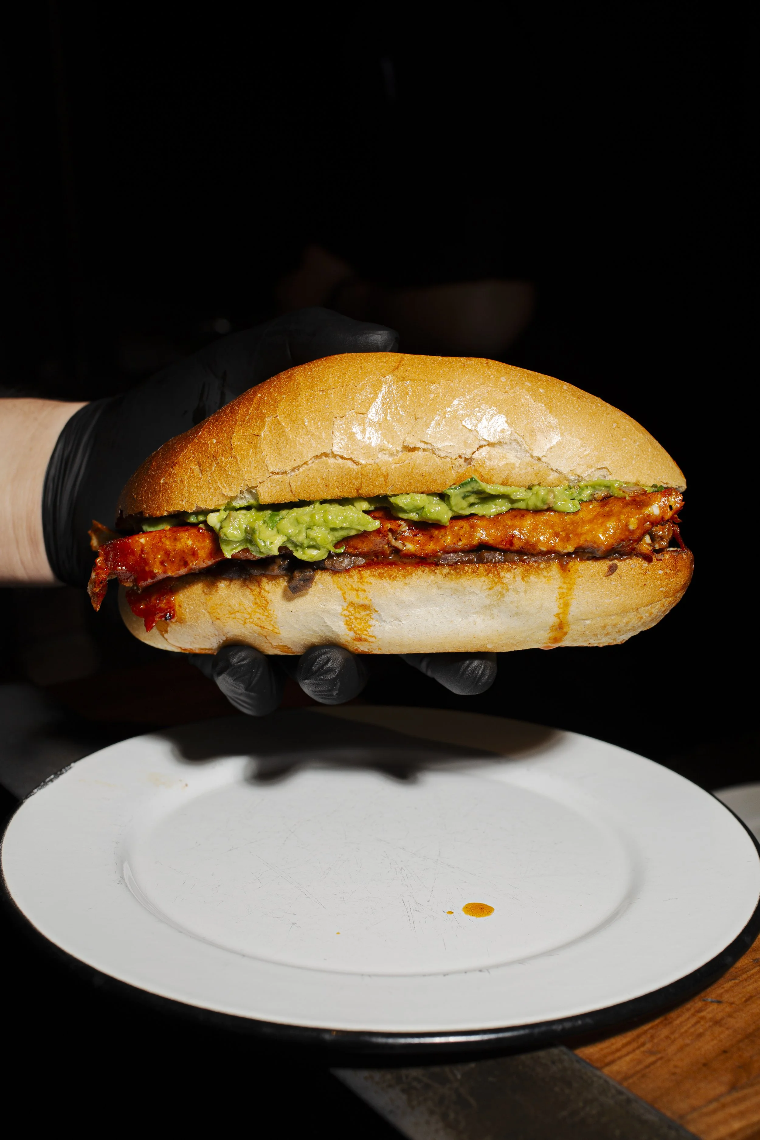 A sandwich with bacon, guacamole, and other ingredients in a soft bun, held by a hand wearing a black glove.