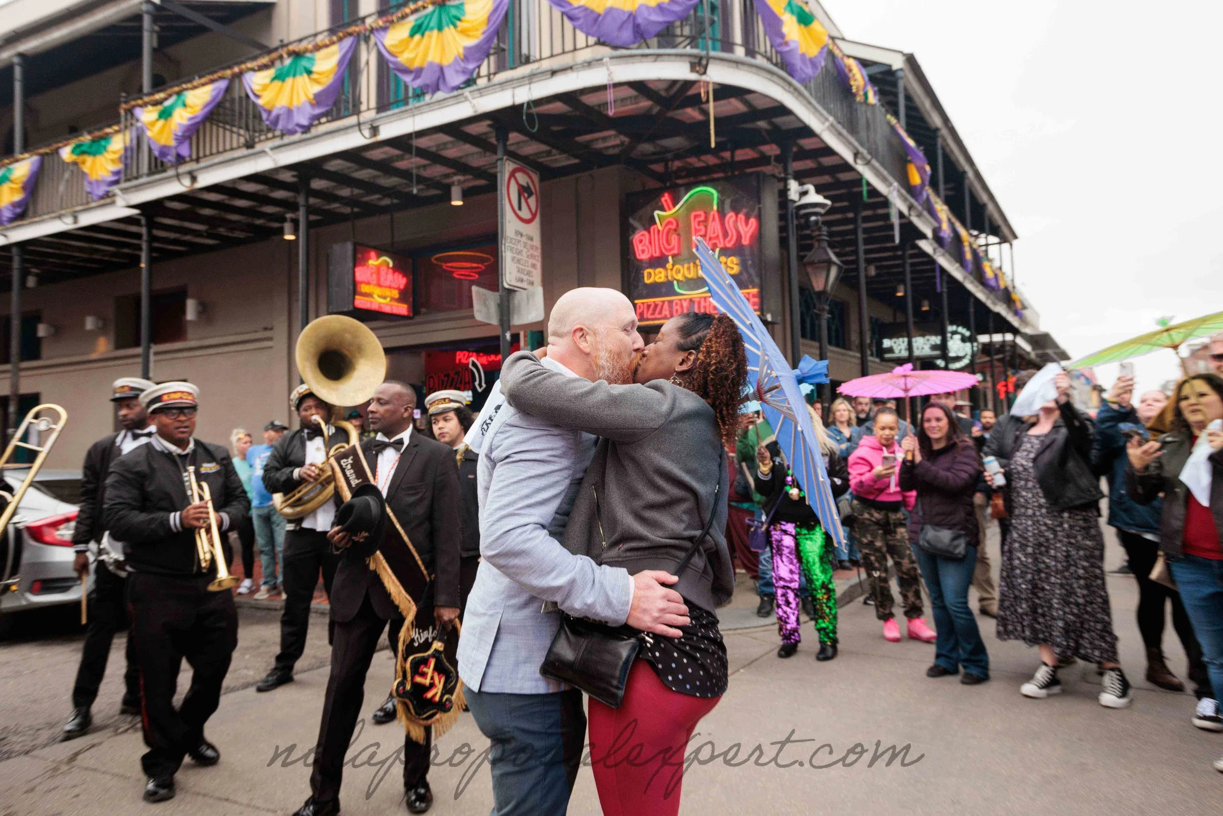 A couple kisses in front of a new orleans brass band following their mardi gras surprise proposal on bourbon street in new orleans.