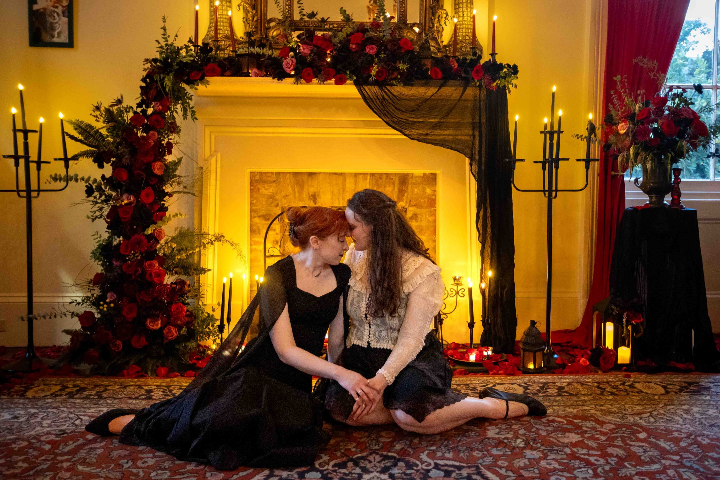 A lesbian couple nestles together in front of a fireplace decorated with gothic romantic decor after their surprise proposal in a New Orleans Mansion.