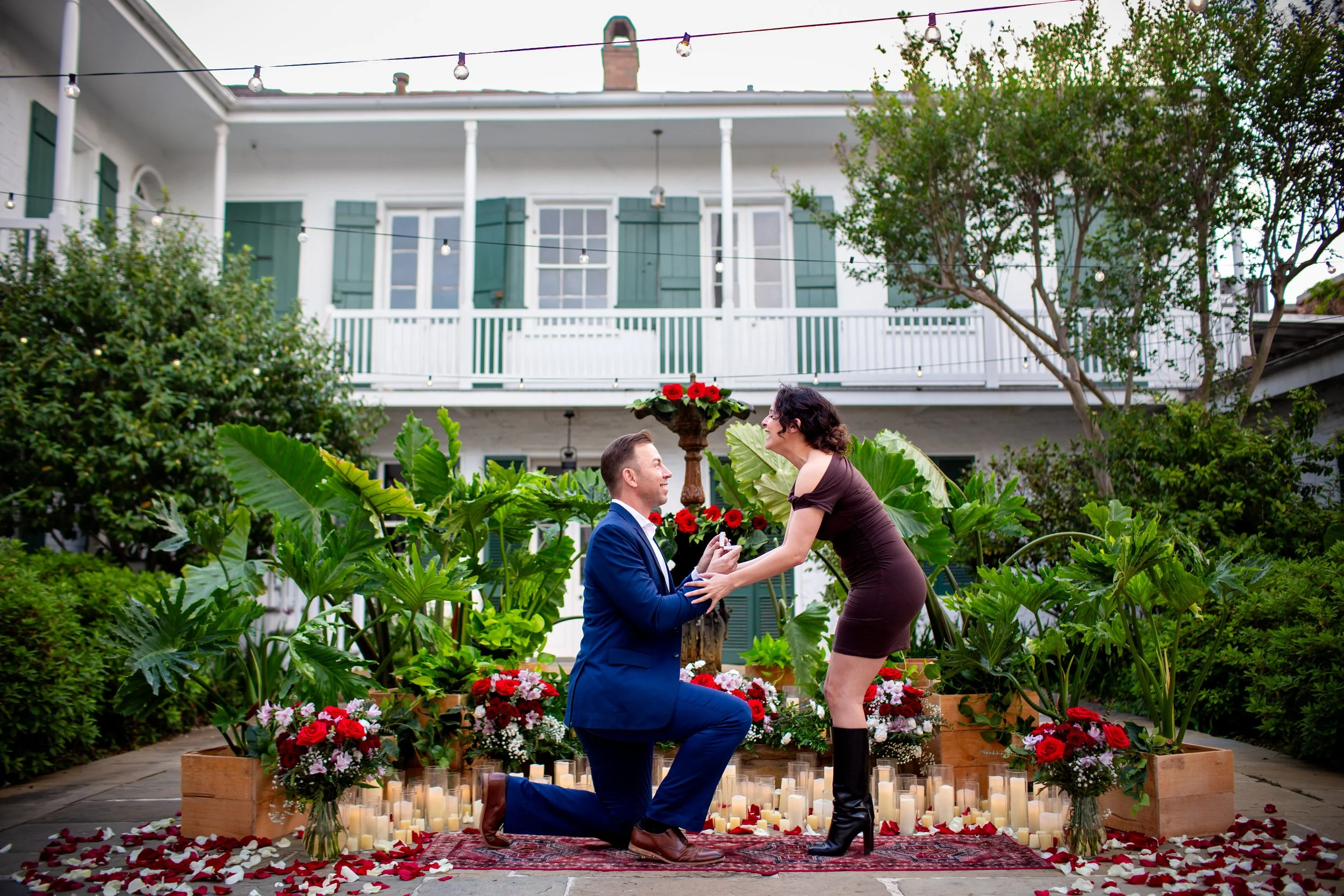 A woman is surprised as her boyfriend is down on one knee proposing in front of a fountain decorated with candles and floral arrangements in a New Orleans French Quarter courtyard.