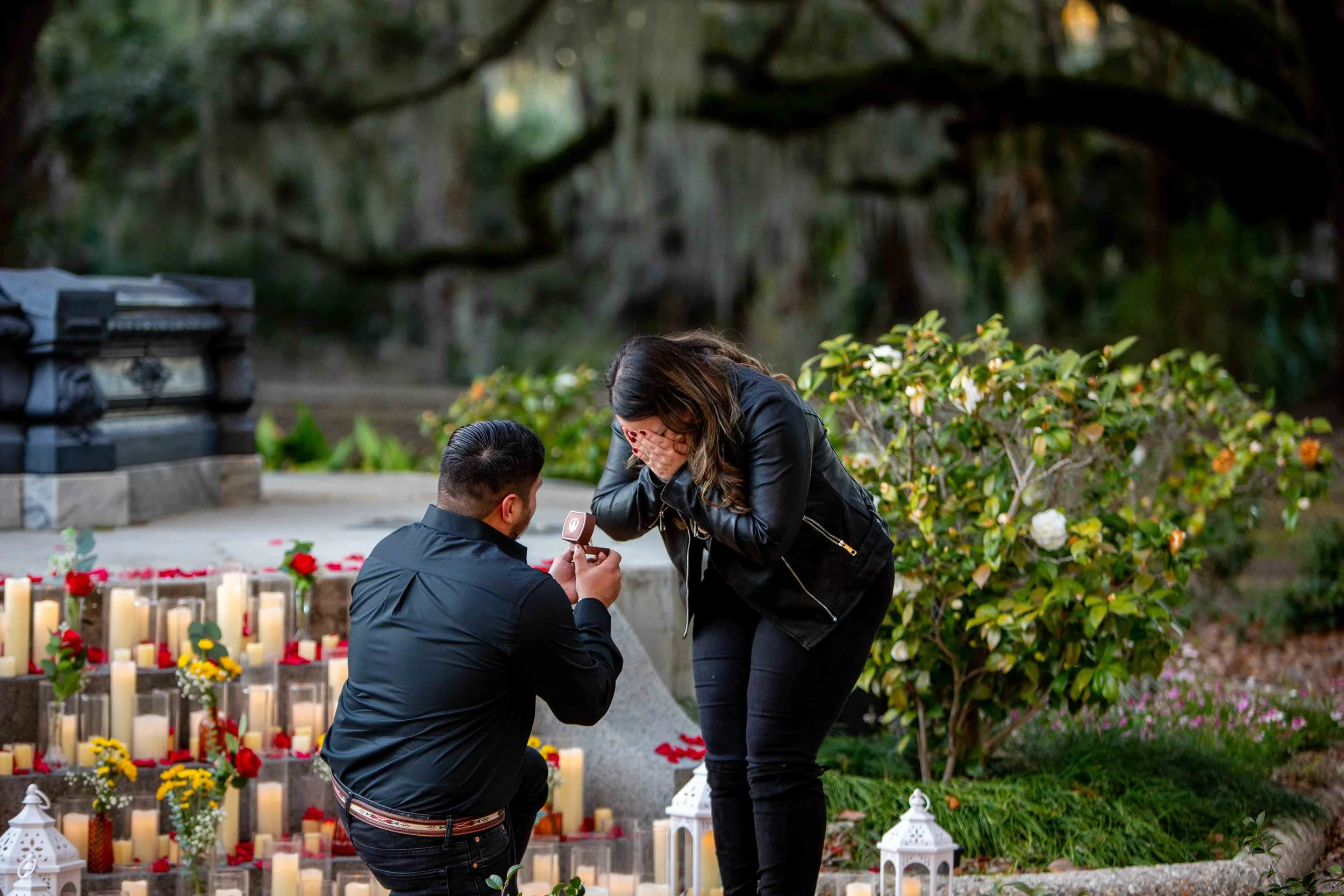 A man proposes to his girlfriend in New Orleans City Park in front of a fountain decorated with candles and red rose petals.